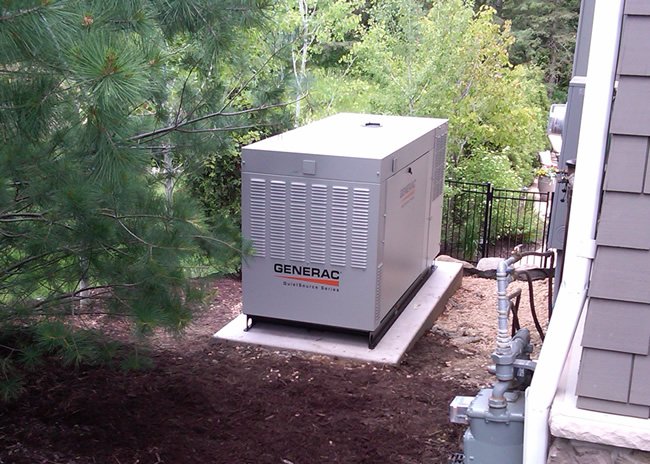 A gray Generac generator on a concrete pad next to a house, surrounded by trees and outdoor equipment.