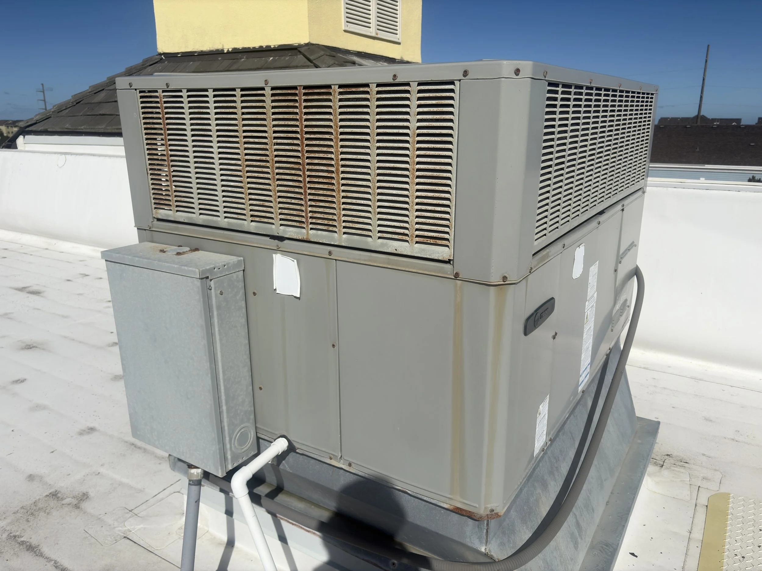 A rooftop air conditioning unit with a rusted front grille and a smaller electrical box attached to its side, with roof and sky in the background.