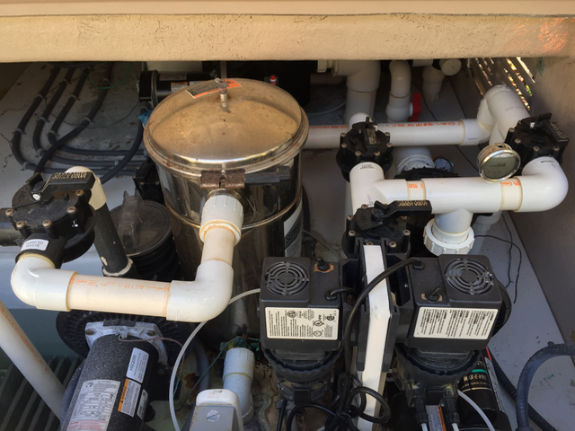 View of a pool or spa pump system with white PVC pipes, pressure gauges, and electric components inside a pump enclosure.