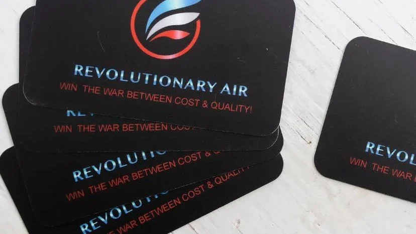 Stack of black coasters with a logo, the text 'REVOLUTIONARY AIR' in blue, and the slogan 'WIN THE WAR BETWEEN COST & QUALITY' in red.