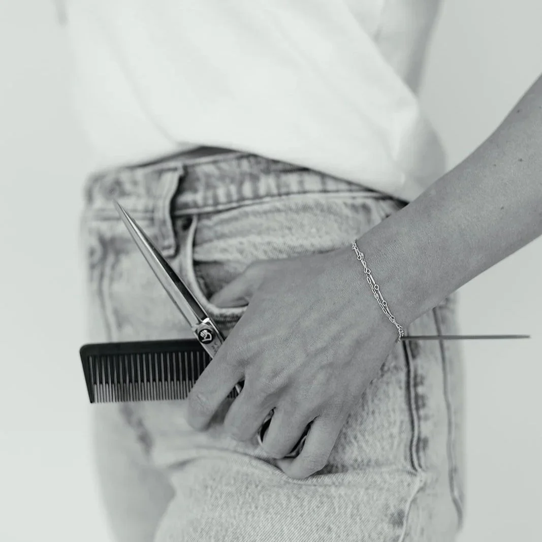 Close-up of a person's hand resting in their jean pocket, holding a black comb and a pair of silver scissors. The person is wearing a thin bracelet on their wrist.