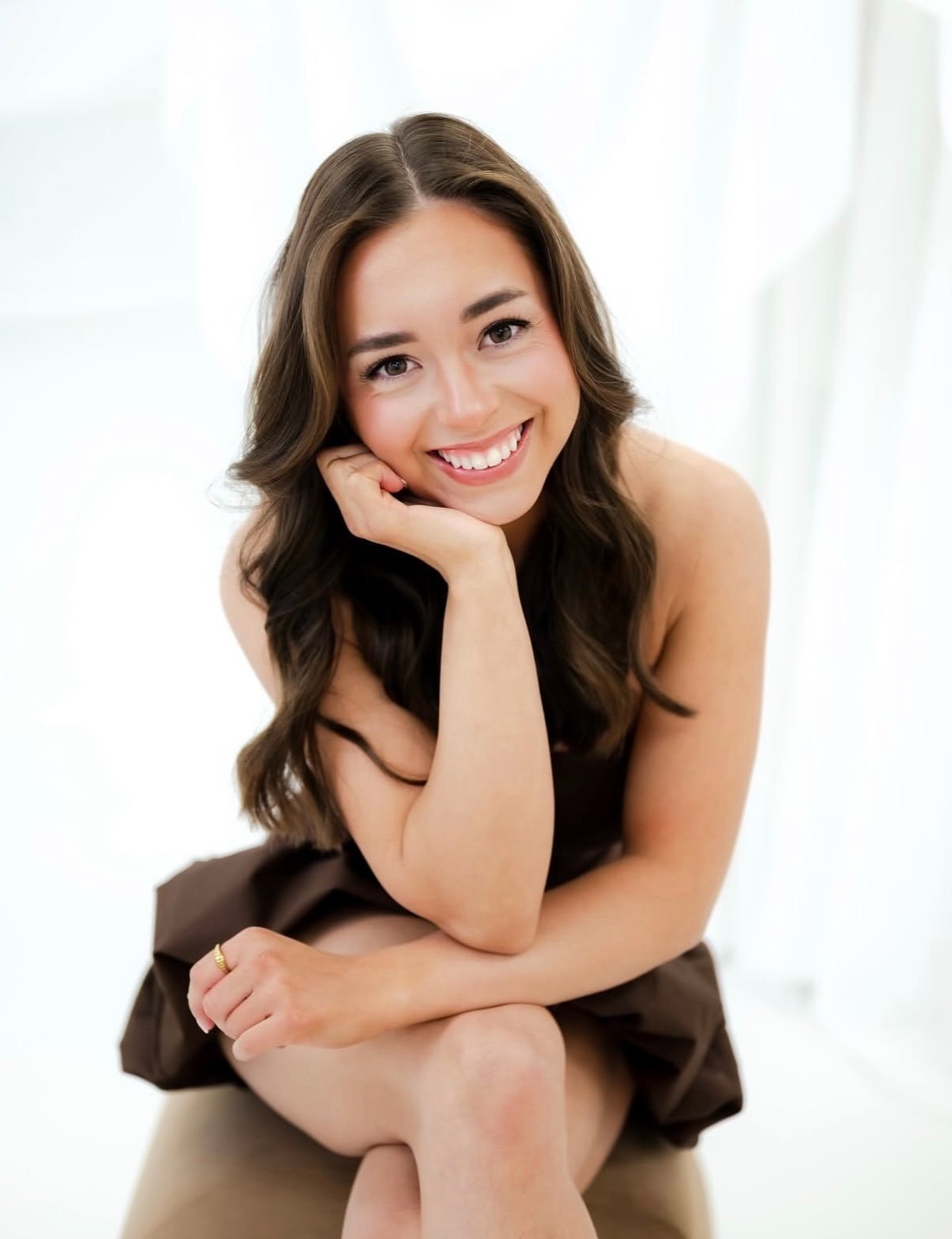 A young woman with brown hair, smiling and resting her chin on her hand, sitting with her legs crossed, against a light background.