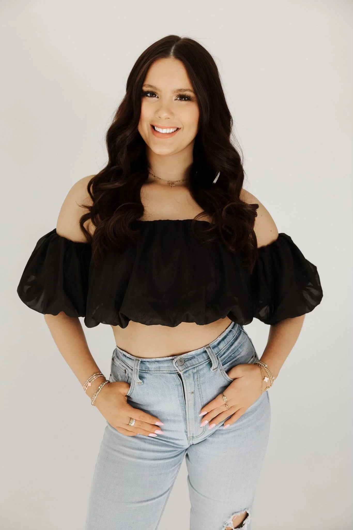 A young woman with brown hair, smiling and resting her chin on her hand, sitting with her legs crossed, against a light background.