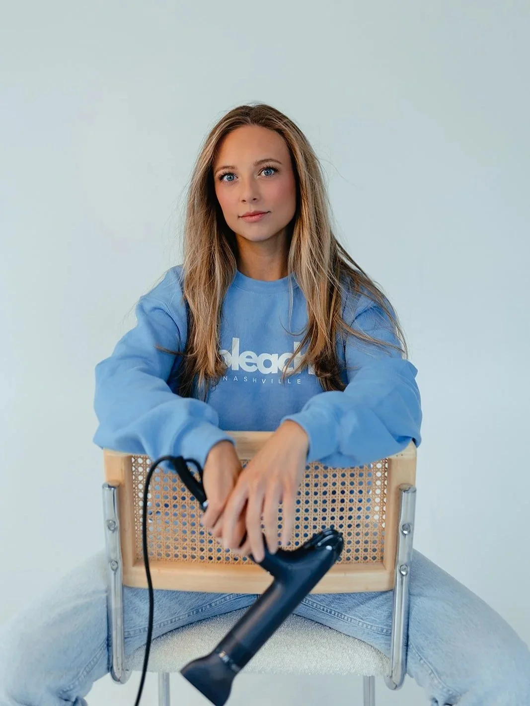 A young woman with long, wavy hair sitting cross-legged on a chair, wearing a light blue sweatshirt with white text, holding a black hairdryer. She is looking directly at the camera with a slight smile, against a plain white background.
