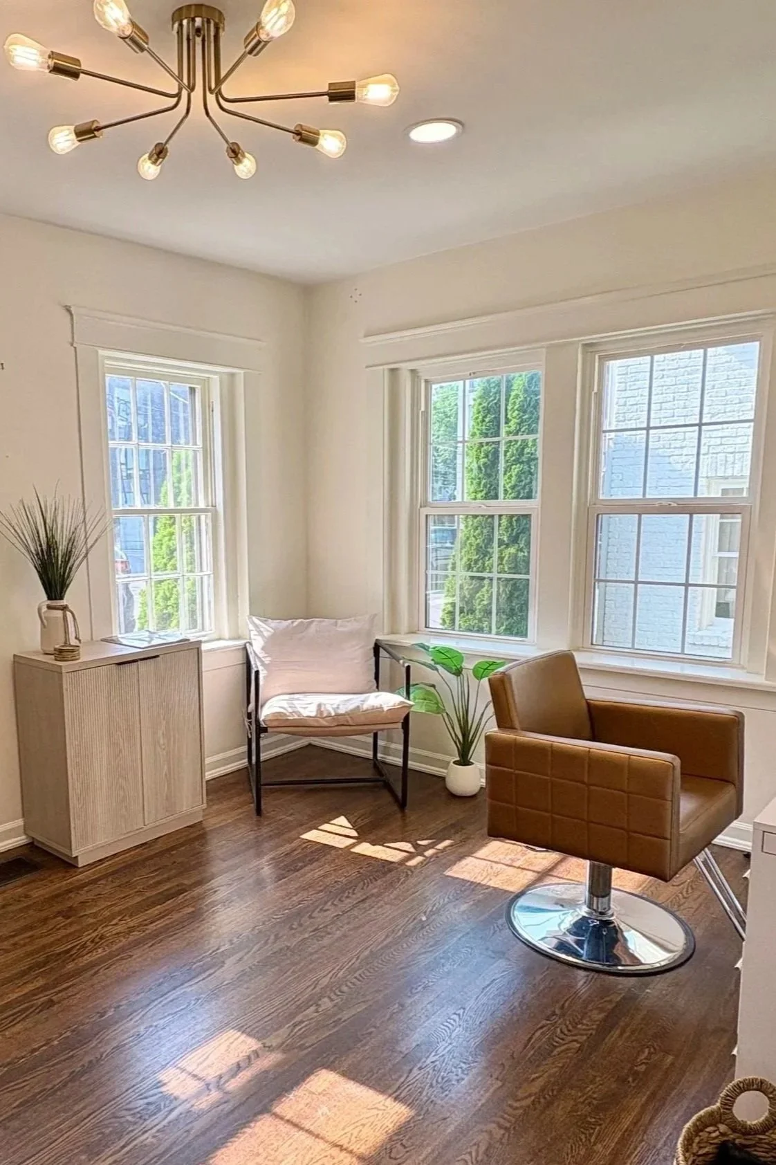 A bright room with white walls and wooden flooring, featuring a modern chandelier on the ceiling, two large windows allowing sunlight, a brown salon chair, a small white side table with a plant, and a black chair with a white cushion near the windows.