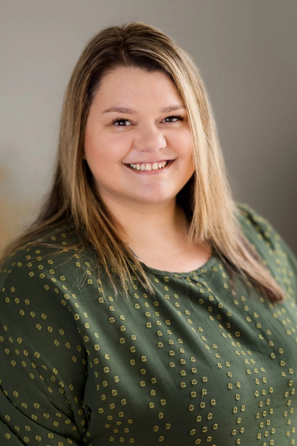 A smiling woman with shoulder-length light brown hair and light skin, wearing a green top with gold polka dots, posing in front of a neutral background.