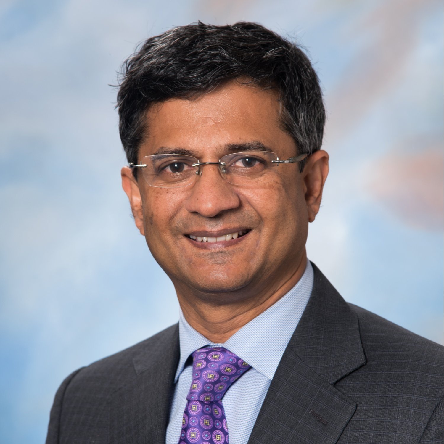 Headshot of a man in a dark suit, light blue shirt, and purple patterned tie, wearing glasses, smiling against a soft cloudy background.
