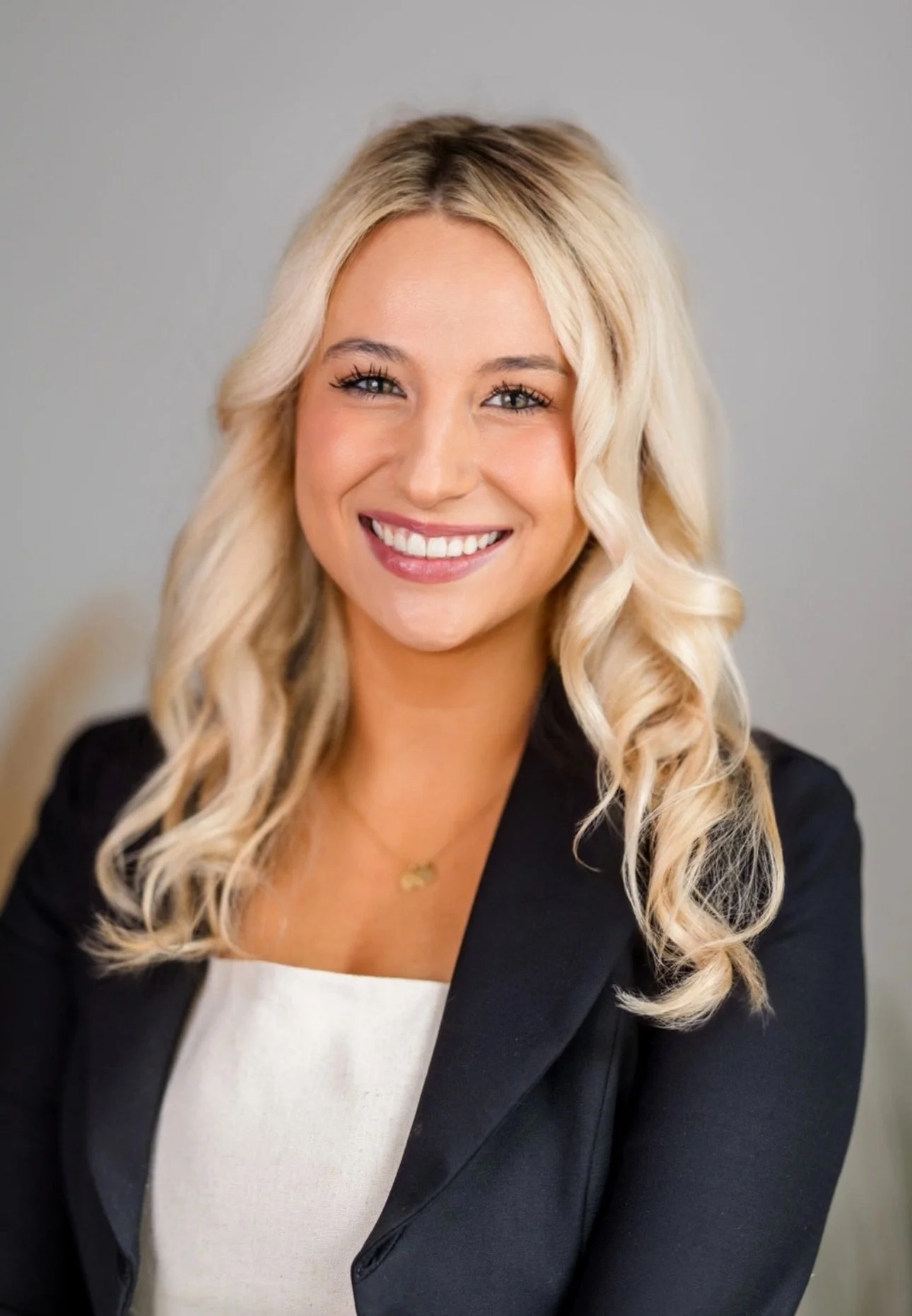 A woman with long, curly blonde hair, wearing a black blazer and white top, smiling against a gray background.