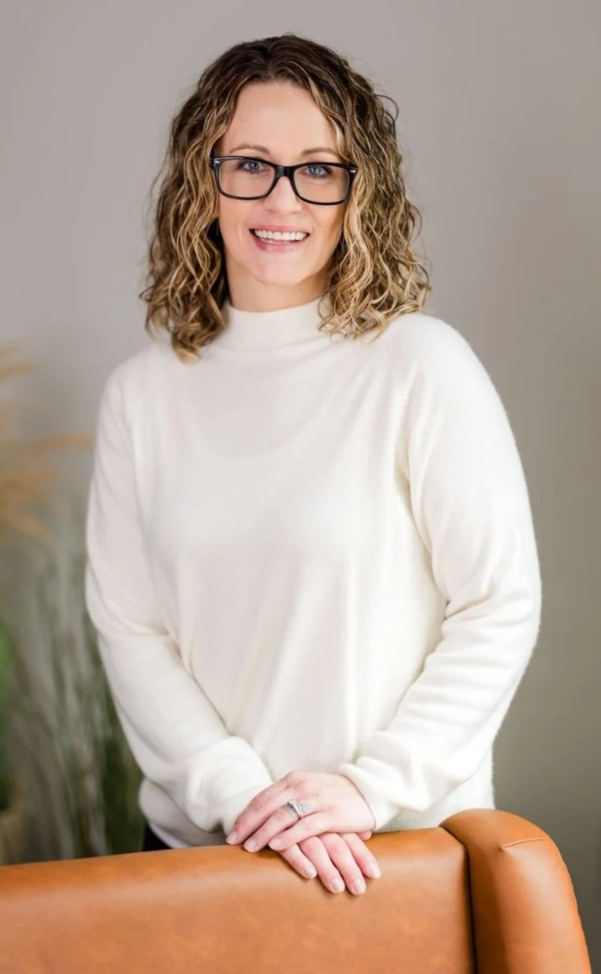 A woman with curly blonde hair, wearing glasses and a white sweater, smiling and leaning on the back of a tan leather chair.