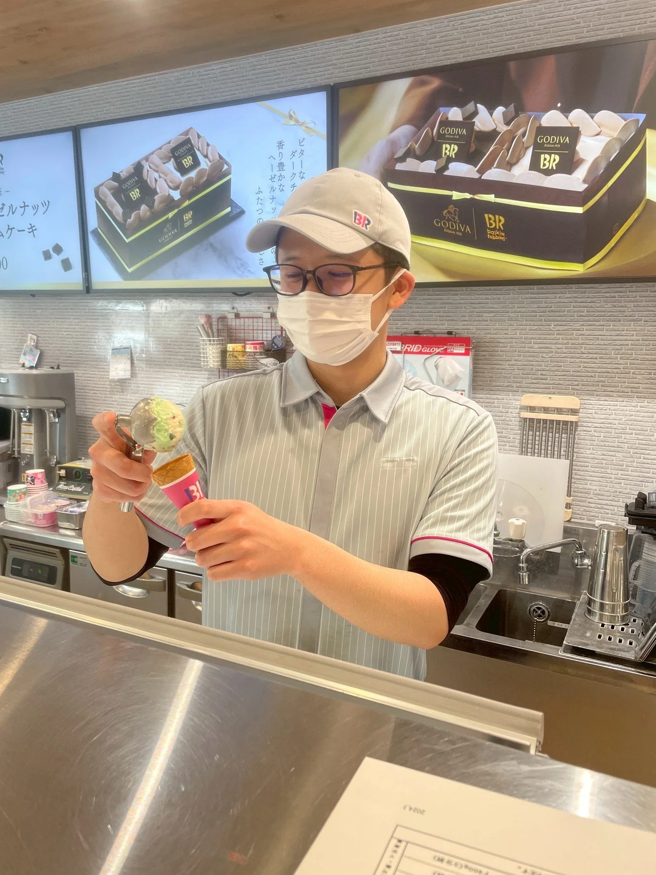 アイスクリームショップで、店員がアイスクリームを仕上げている様子。店員はマスクと帽子を着用し、バックに商品の広告が見える。