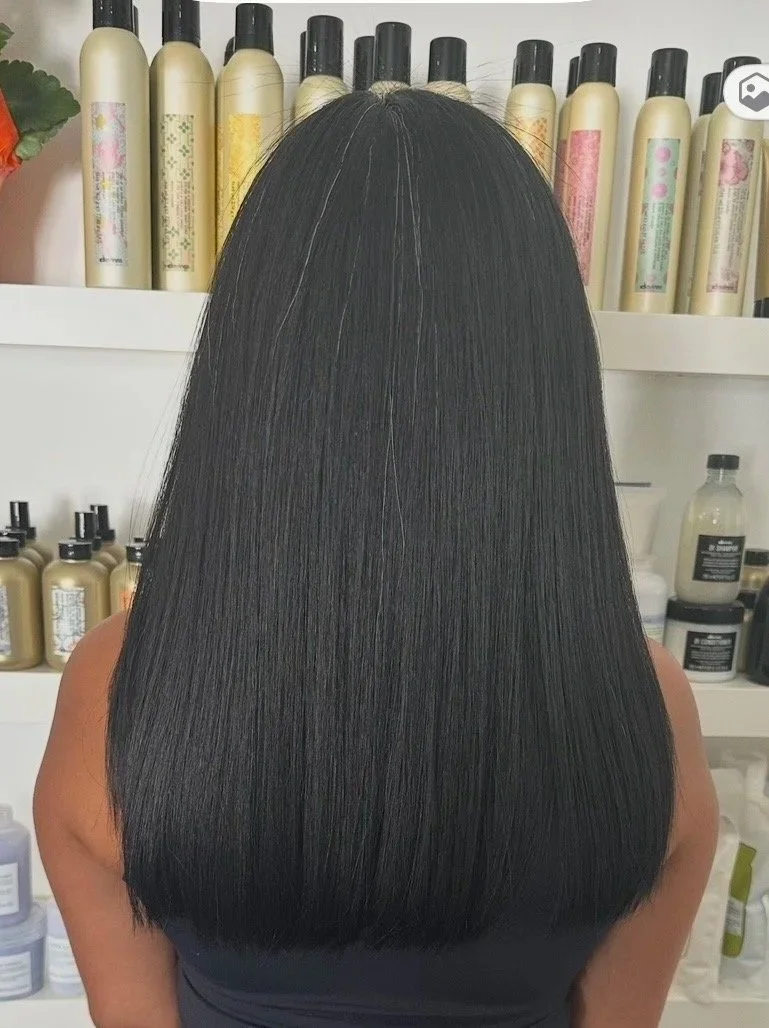 Back view of a woman with long, straight, shiny black hair with some lighter streaks, standing in front of shelves filled with hair care products.