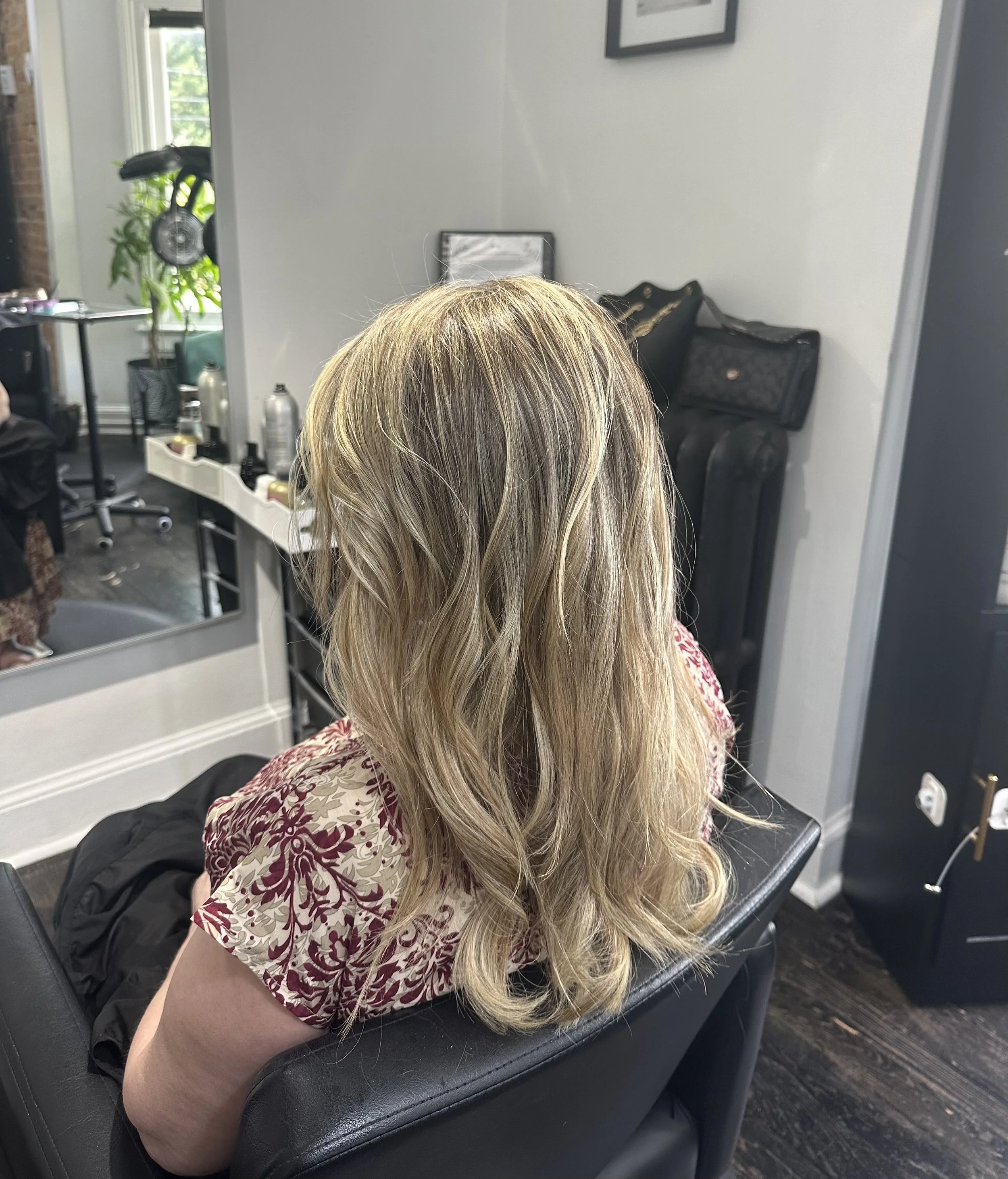 A woman with long blonde hair sitting in a salon chair, wearing a patterned shirt. The salon has a mirror, shelving with styling products, a window with greenery outside, and black bags hanging on the wall.
