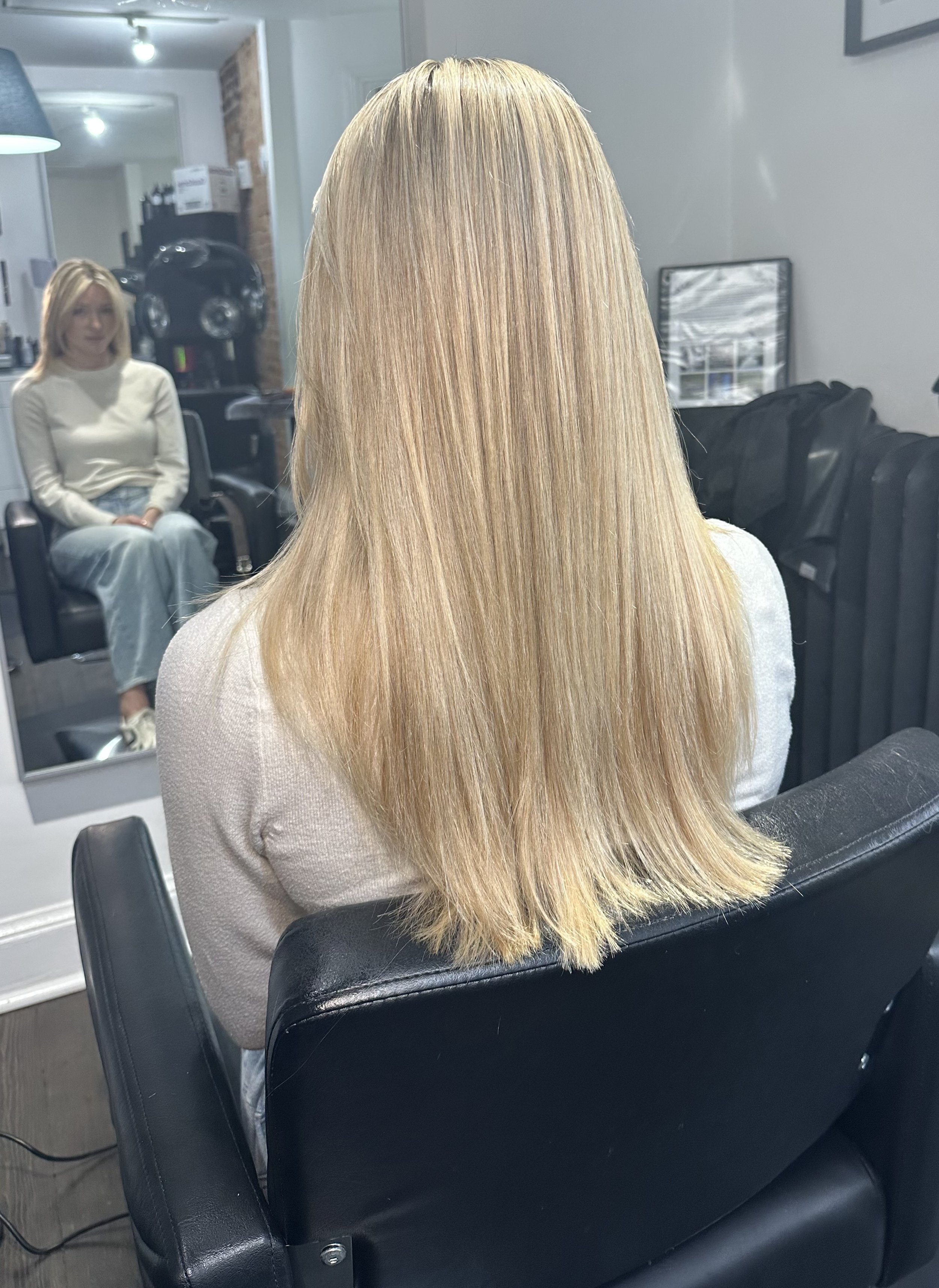A person with long, straight, blonde hair sitting in a black salon chair, looking into a mirror, with a woman with shoulder-length blonde hair seated in the background.