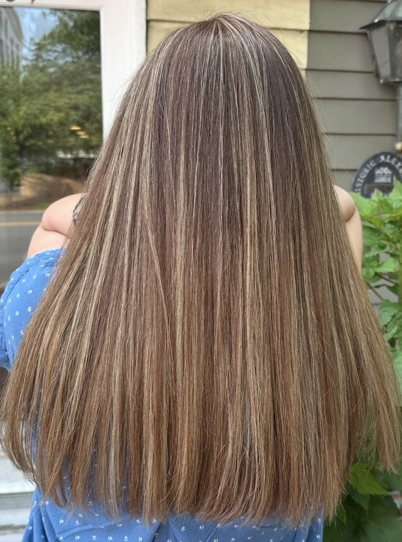 Back of a woman with long, straight, light brown hair with subtle highlights, standing outdoors near a house with beige siding and plants.