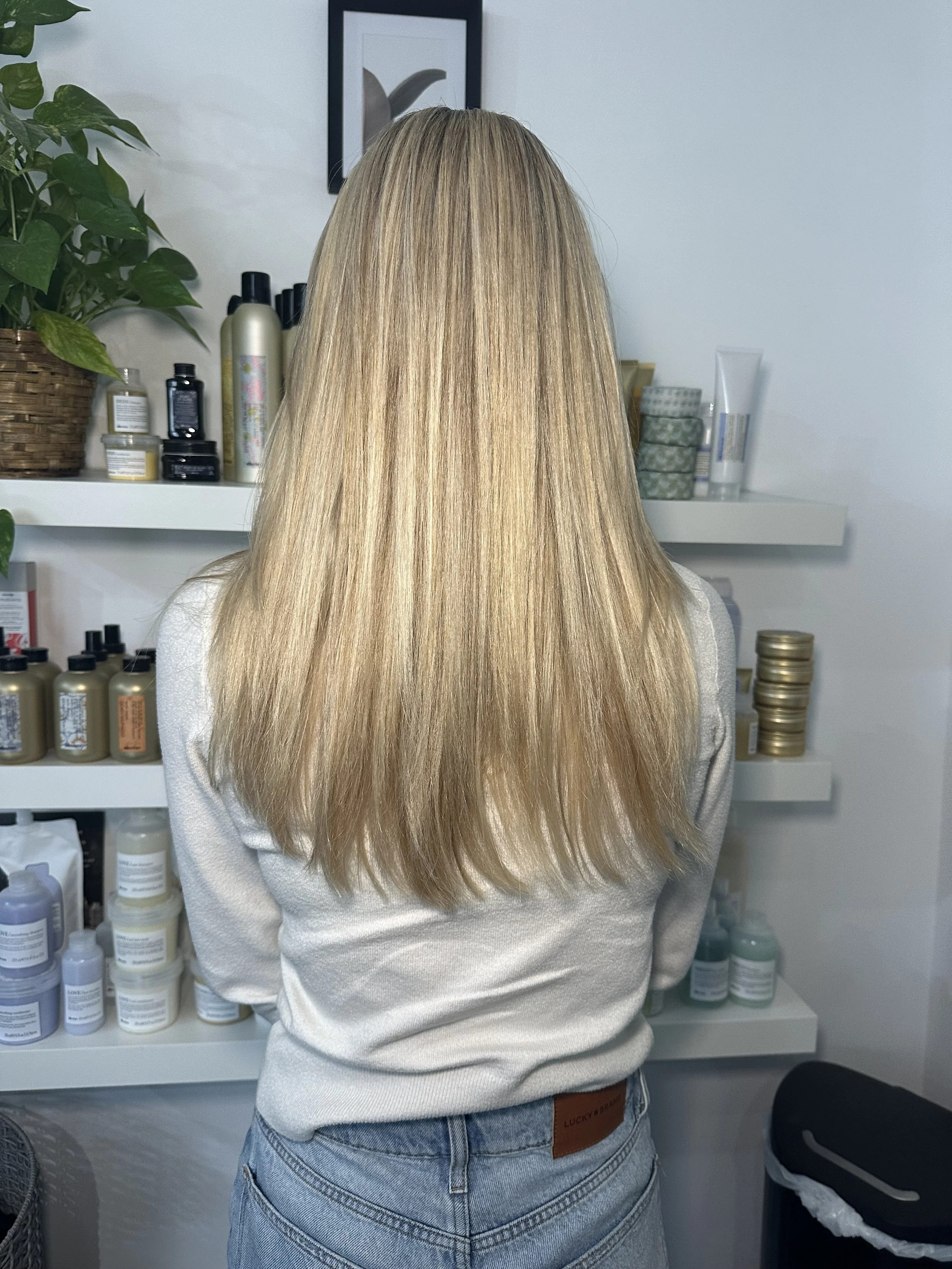 Back view of a woman with long, straight blonde hair wearing a light-colored sweater and jeans, standing in front of shelves with hair and beauty products in a salon or spa.