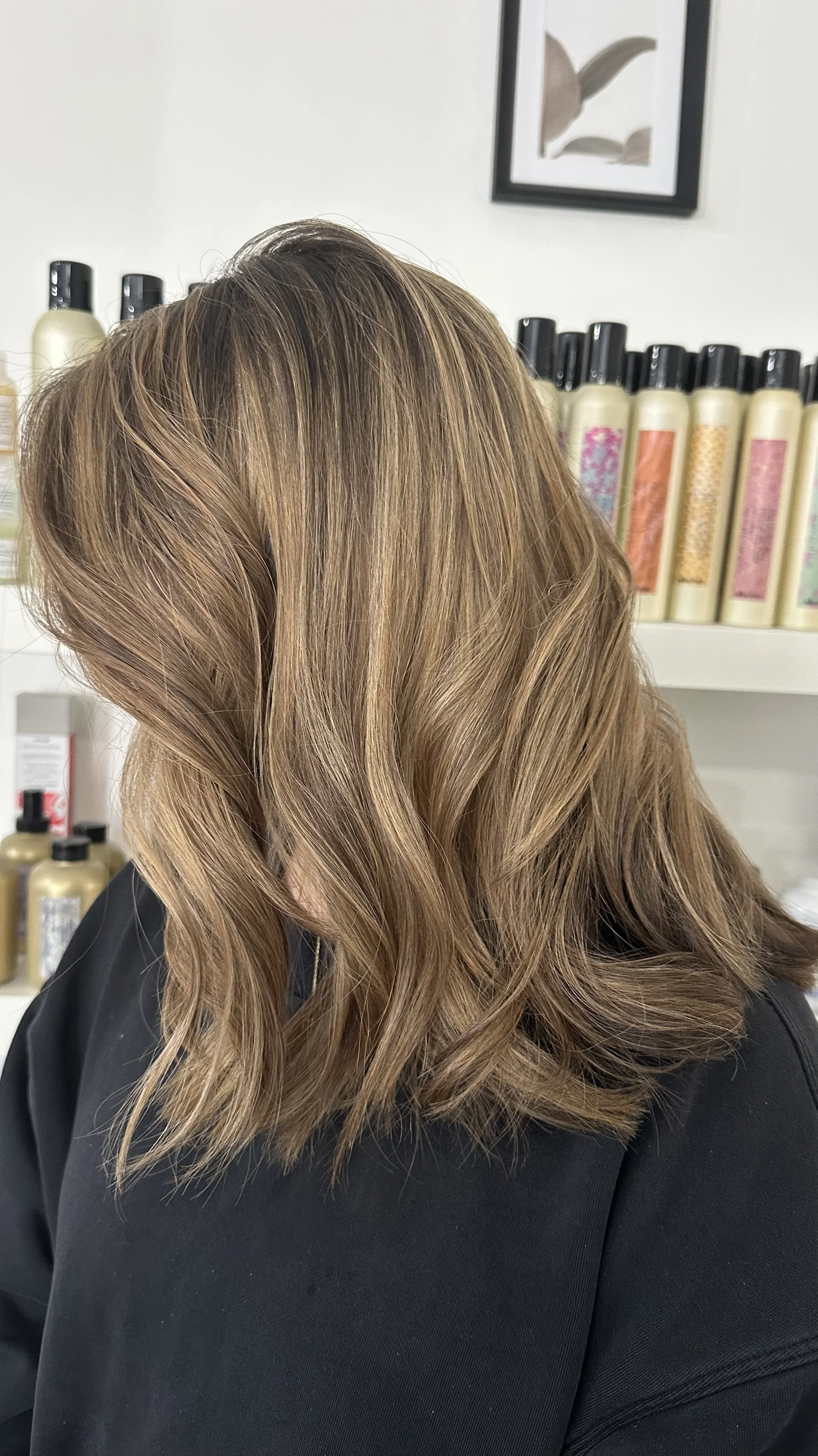 A woman with long, wavy, light brown hair with highlights, sitting in a salon with hair products on shelves in the background.