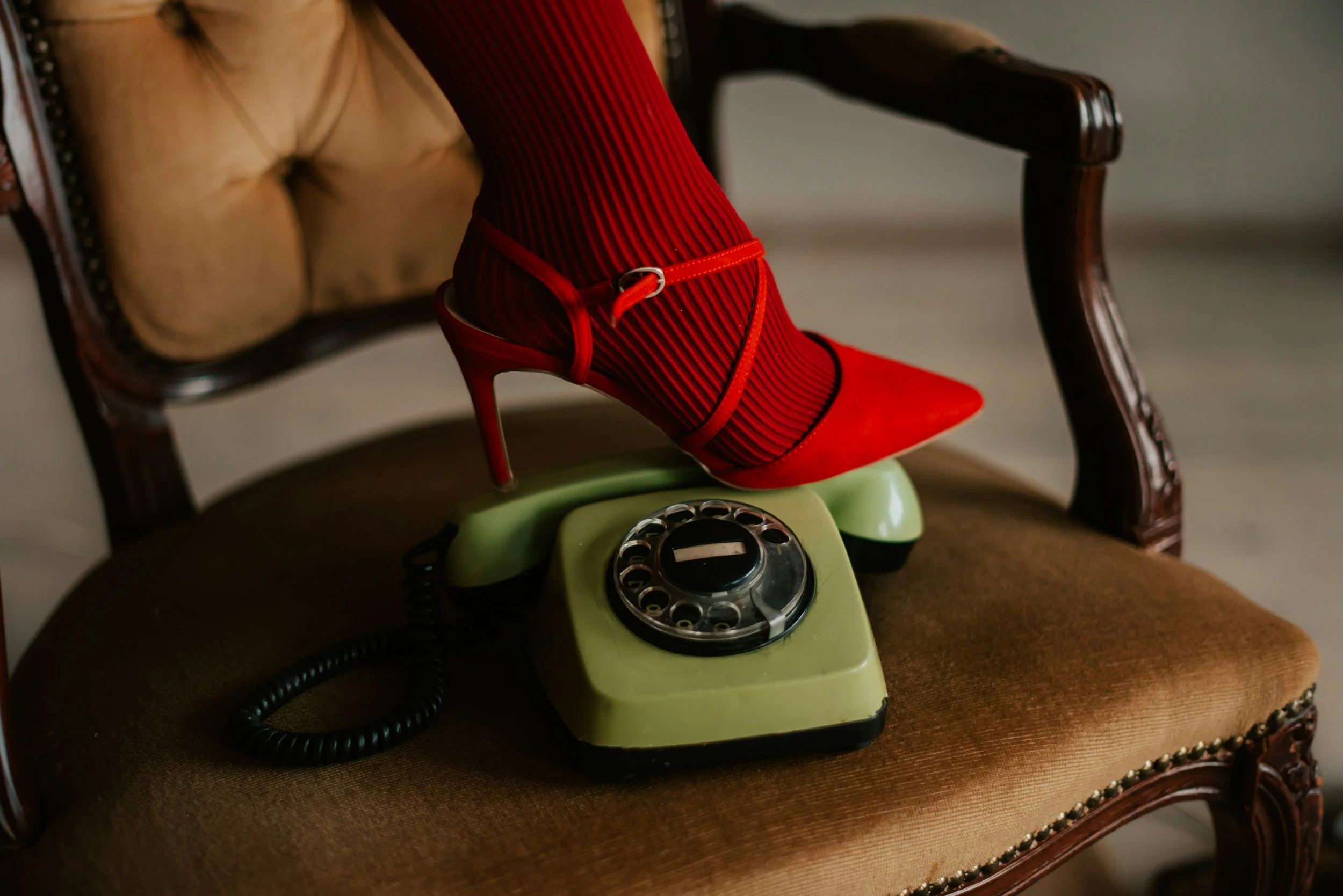 A vintage green rotary phone with a red high-heeled shoe on top and red striped stockings covering the foot, all placed on a brown upholstered chair with wooden armrests.