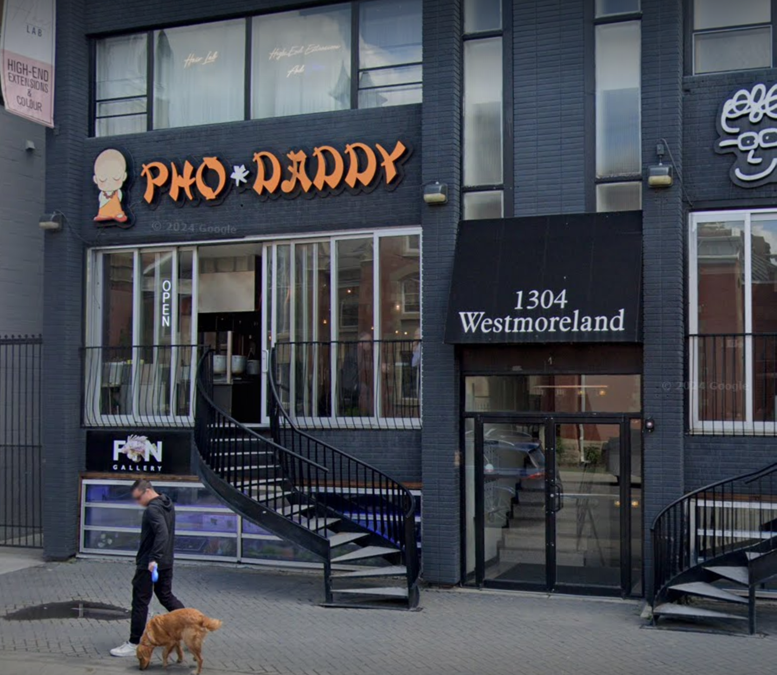 Exterior of a modern black brick building with large glass windows and a black sign that reads '1304 Westmoreland.' There are two spiral staircases, one on the left and one on the right. A man is walking a brown dog on the sidewalk in front of the building, and a person is seen inside through the windows.