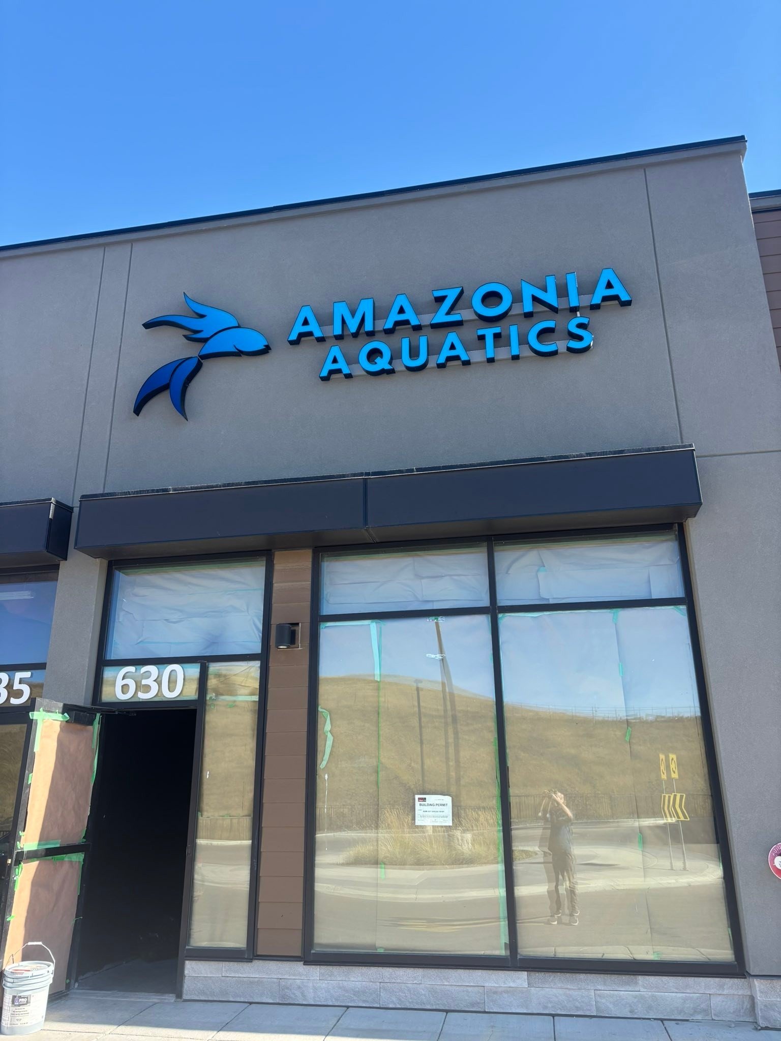 Storefront with a sign that reads 'Amazonia Aquatics' and a stylized fish logo, with large glass windows and a door, under a clear blue sky.