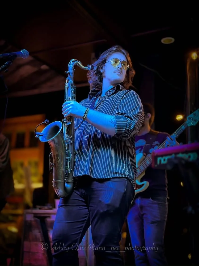 A person with shoulder-length hair wearing sunglasses and a striped shirt playing the saxophone on stage with another musician in the background playing the guitar.