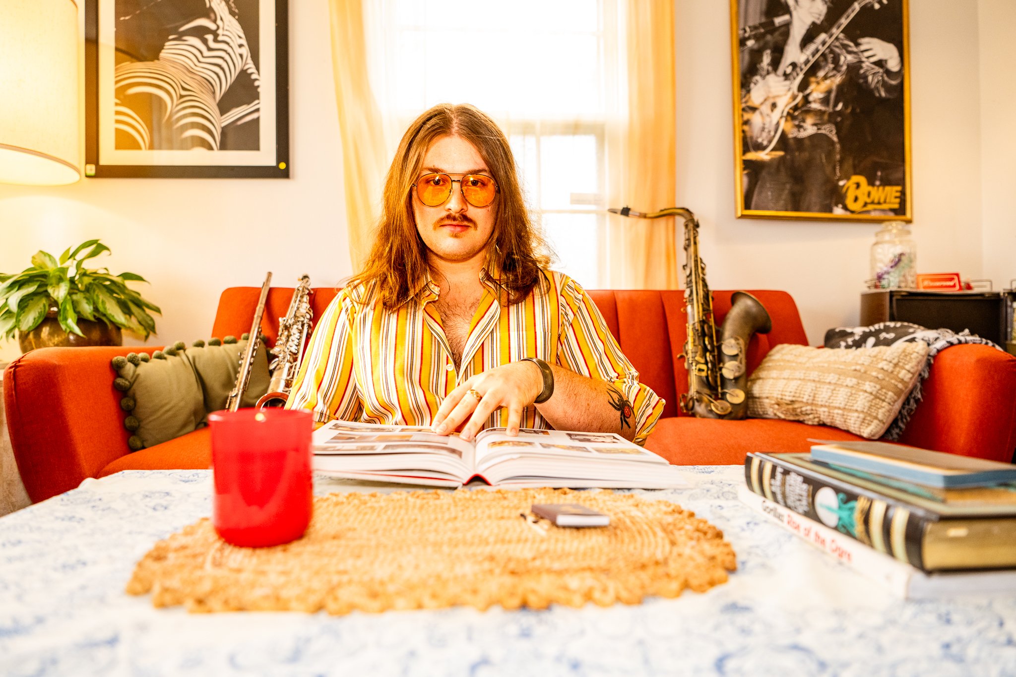 A man with long hair, wearing orange-tinted sunglasses and a striped yellow, orange, and white shirt, sits at a table with a photo album open. Behind him on a red couch are musical instruments, including a saxophone and a guitar. The room has posters on the wall, including one of David Bowie, and a plant on the left side.