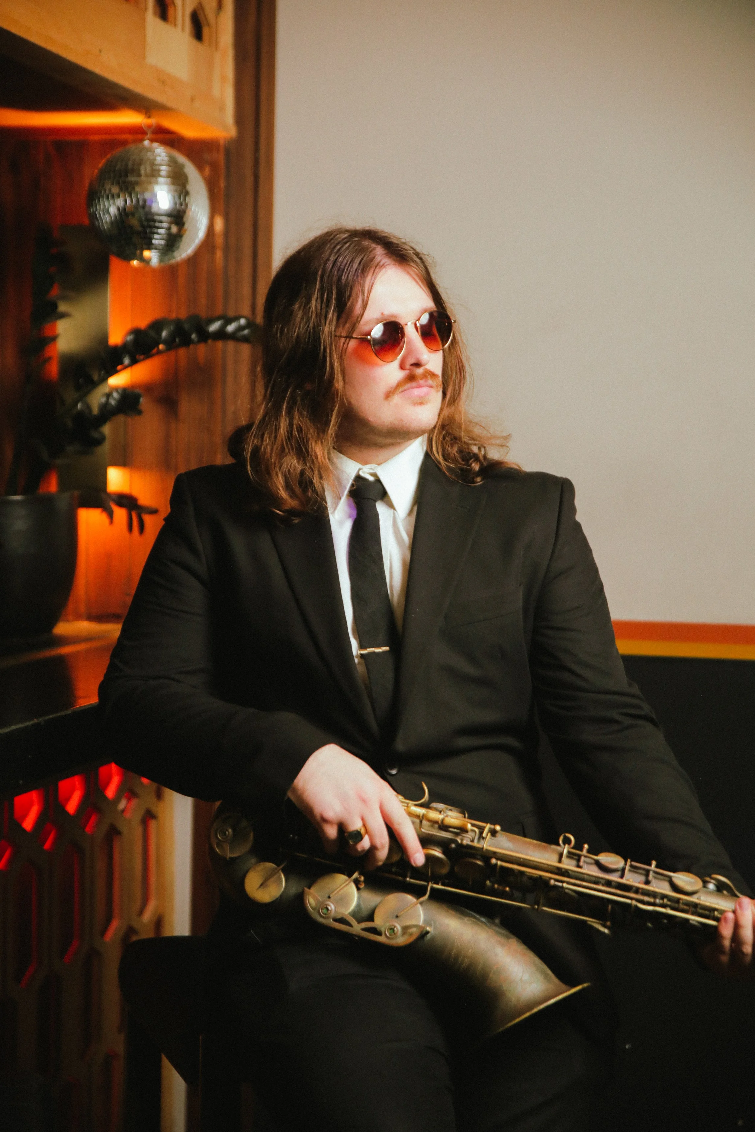 A man with long hair, sunglasses, and a mustache dressed in a black suit and tie, sitting with a saxophone in hand in a retro-style setting.