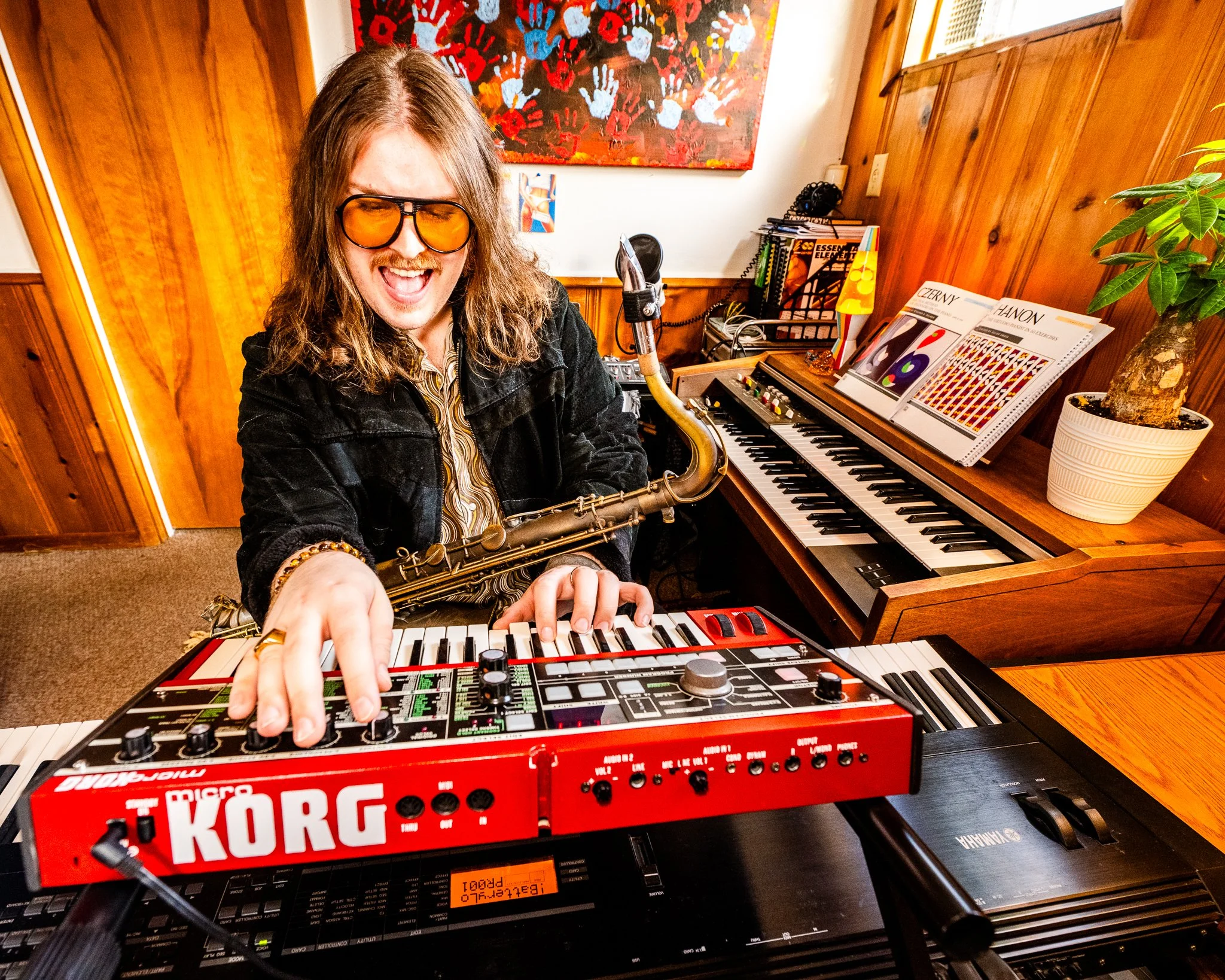 A man with long hair, wearing glasses and a black jacket, playing a red Korg synthesizer keyboard while smiling. He is in a music room with wooden walls, a piano with books and sheet music, and colorful artwork in the background.