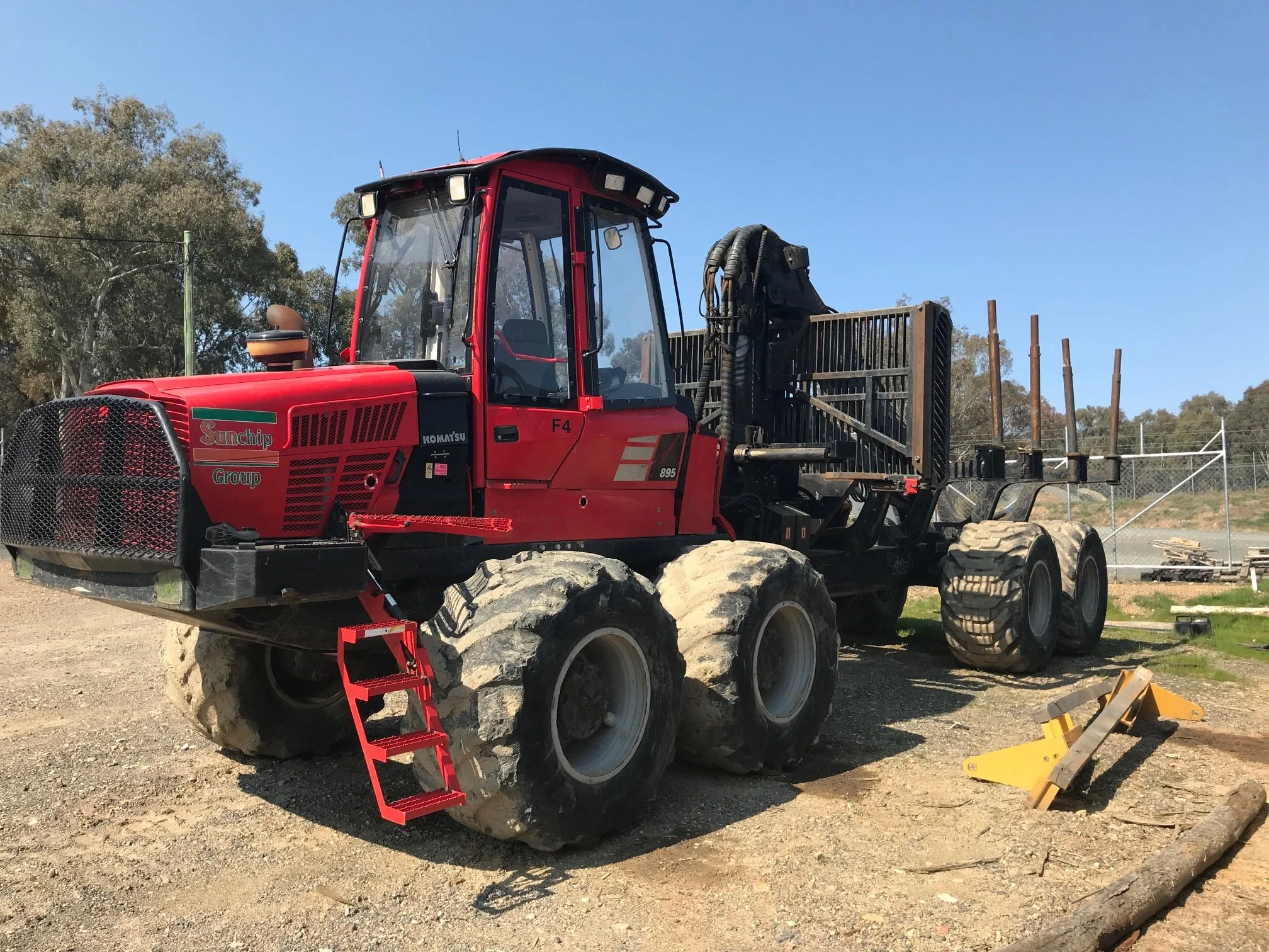 Red and black forestry machine parked on a dirt surface with large tires, a protective grill, and cabin, surrounded by trees and a clear blue sky.