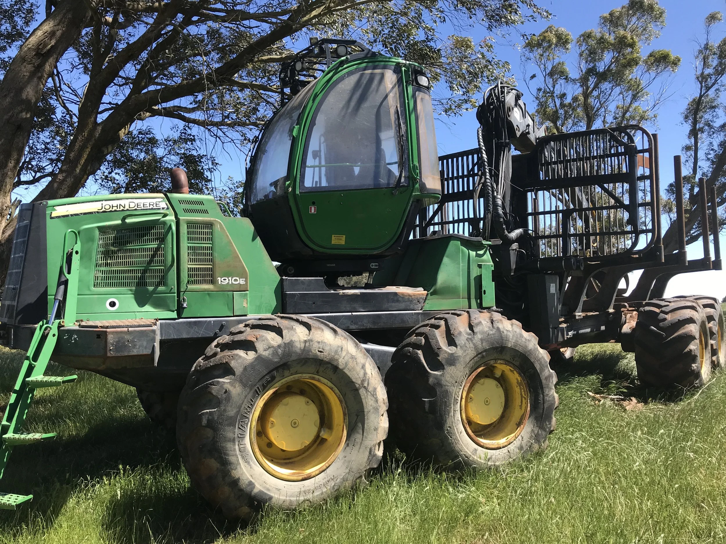 A green John Deere forestry forwarder with large tires, parked on grass under trees.