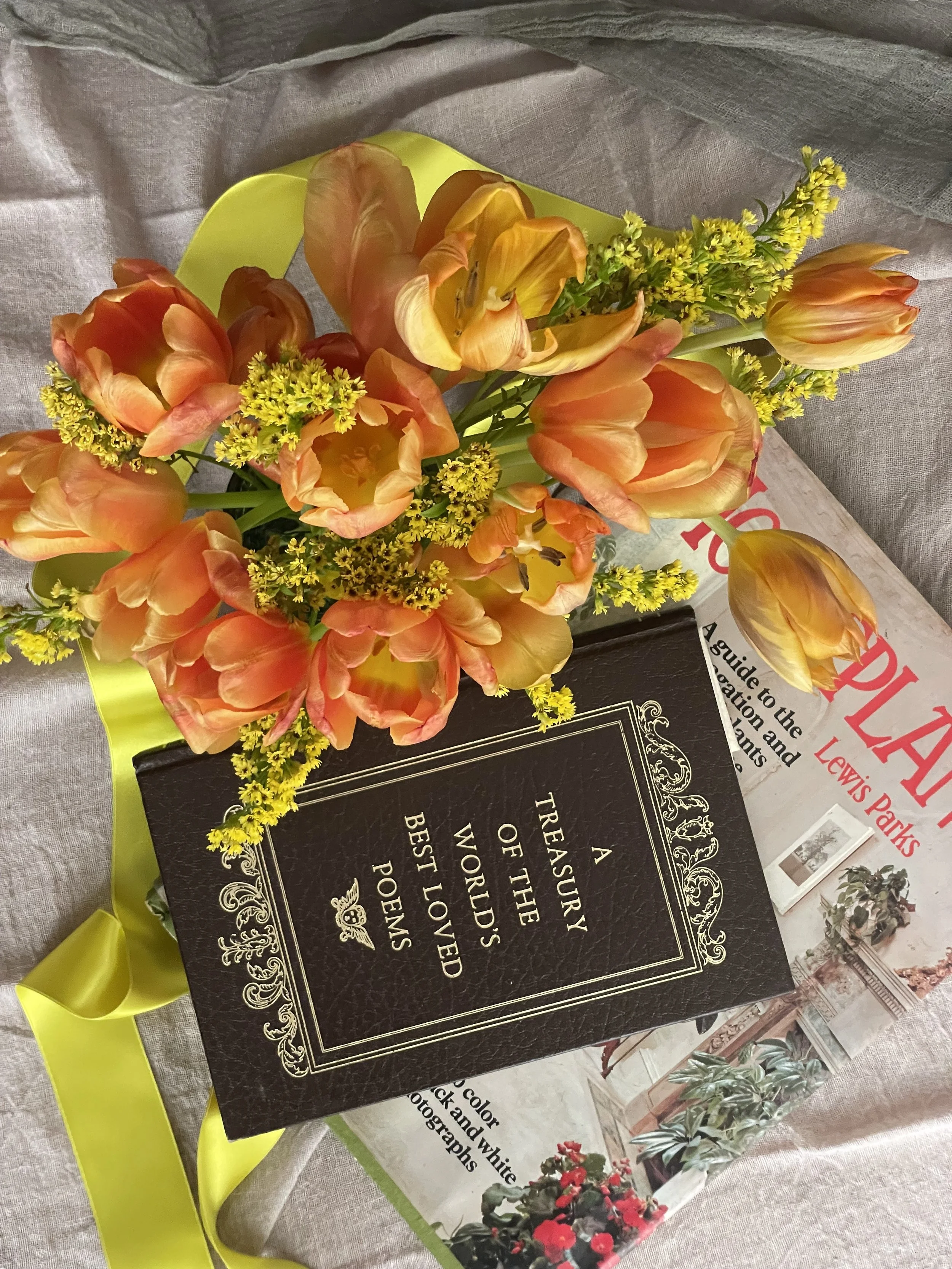 A bouquet of orange tulips and yellow flowers resting on a black book titled 'A Treasure of the World's Best Loved Poems' with a yellow ribbon and a magazine in the background.