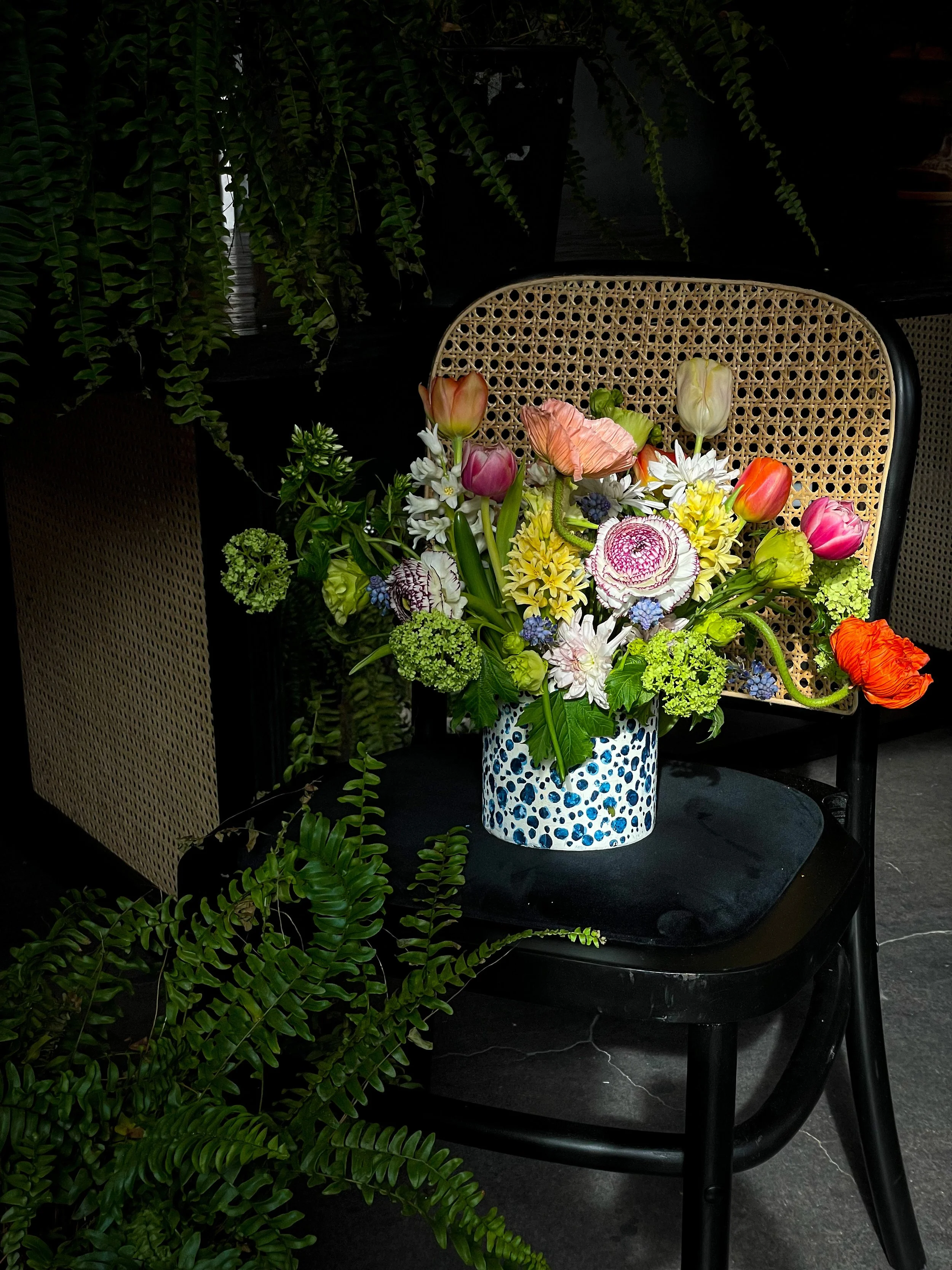 A colorful bouquet of flowers in a patterned vase on a black chair with green plants in the background.