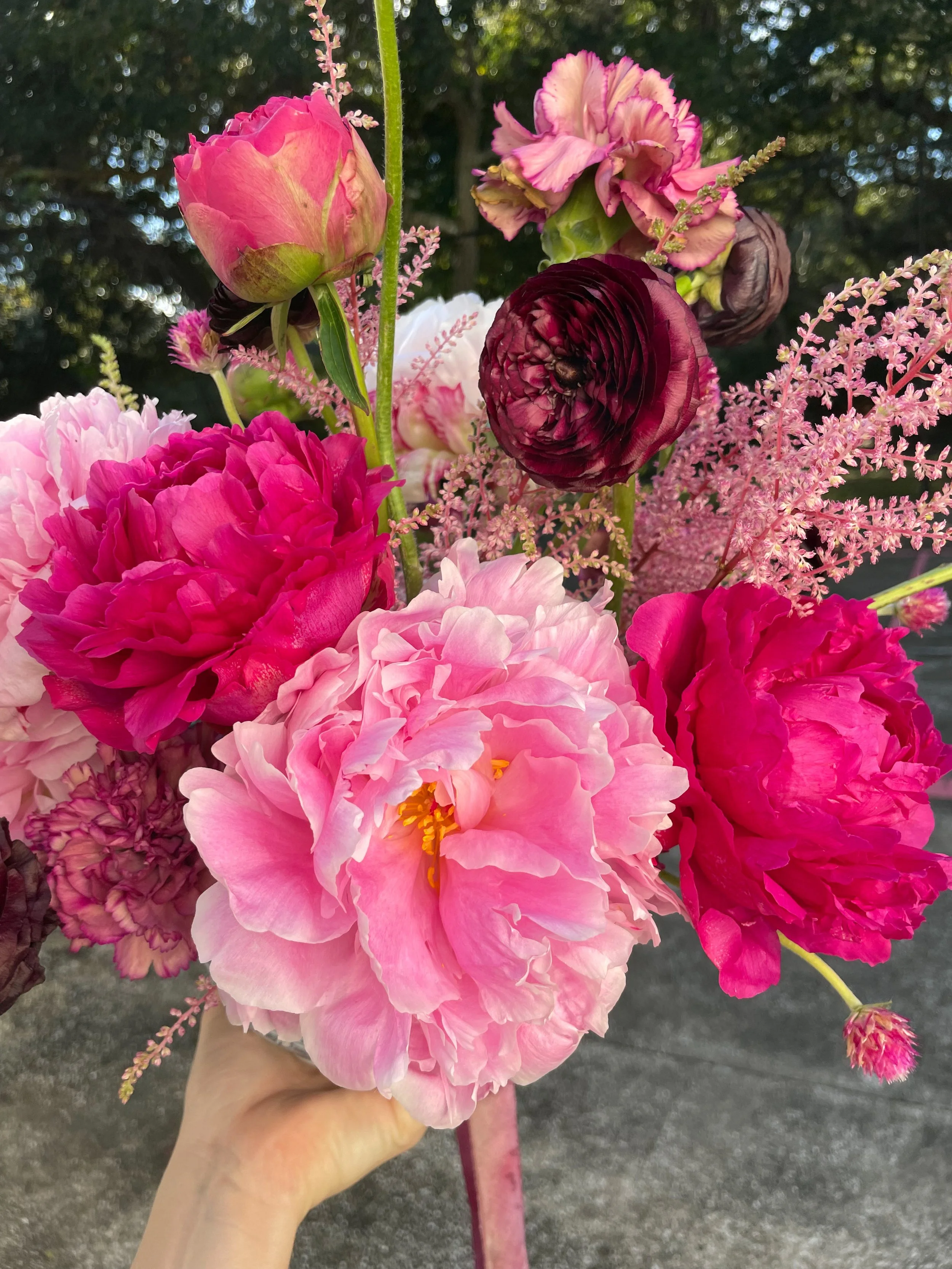 A colorful bouquet of pink, purple, and red peonies with delicate pink astilbe flowers on a green stem, held by a person's hand, outdoors with trees in the background.
