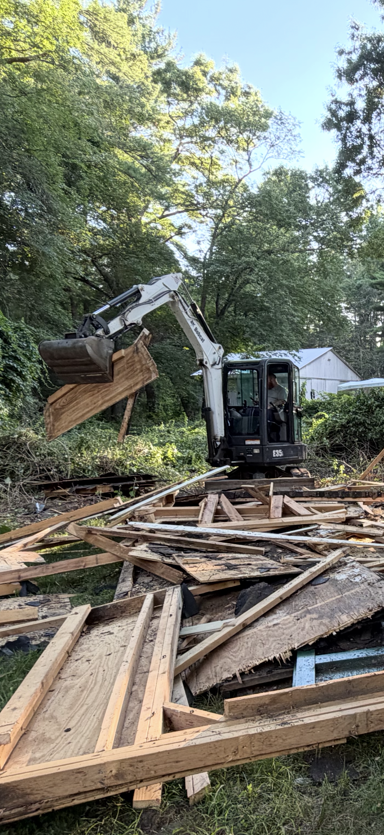 A small excavator is demolishing a wooden structure, with debris and wood scattered on the ground. Trees and a white building are in the background.