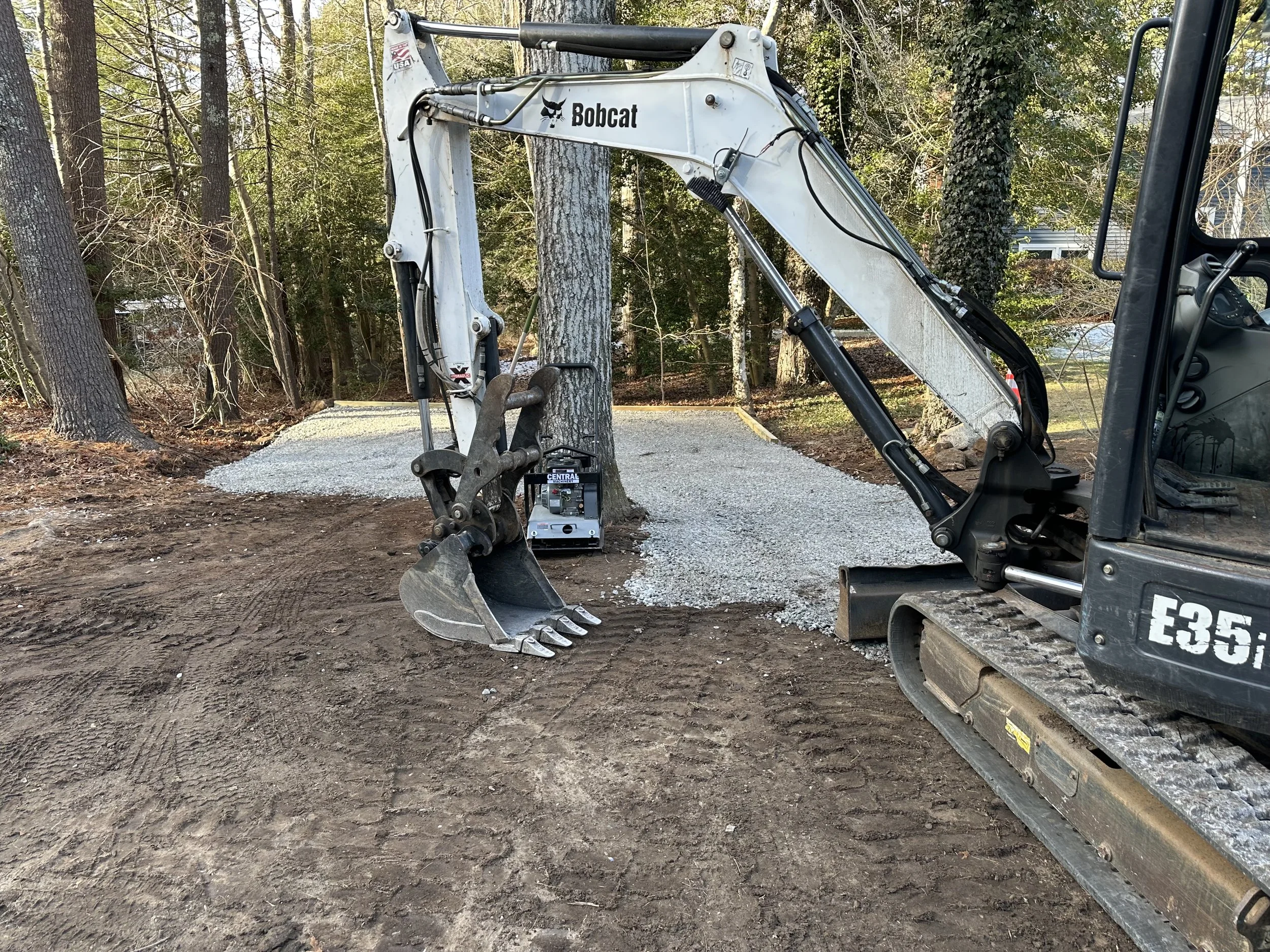 Construction site with a mini excavator labeled 'Bobcat' digging near a tree, gravel path, and wooded area in the background.