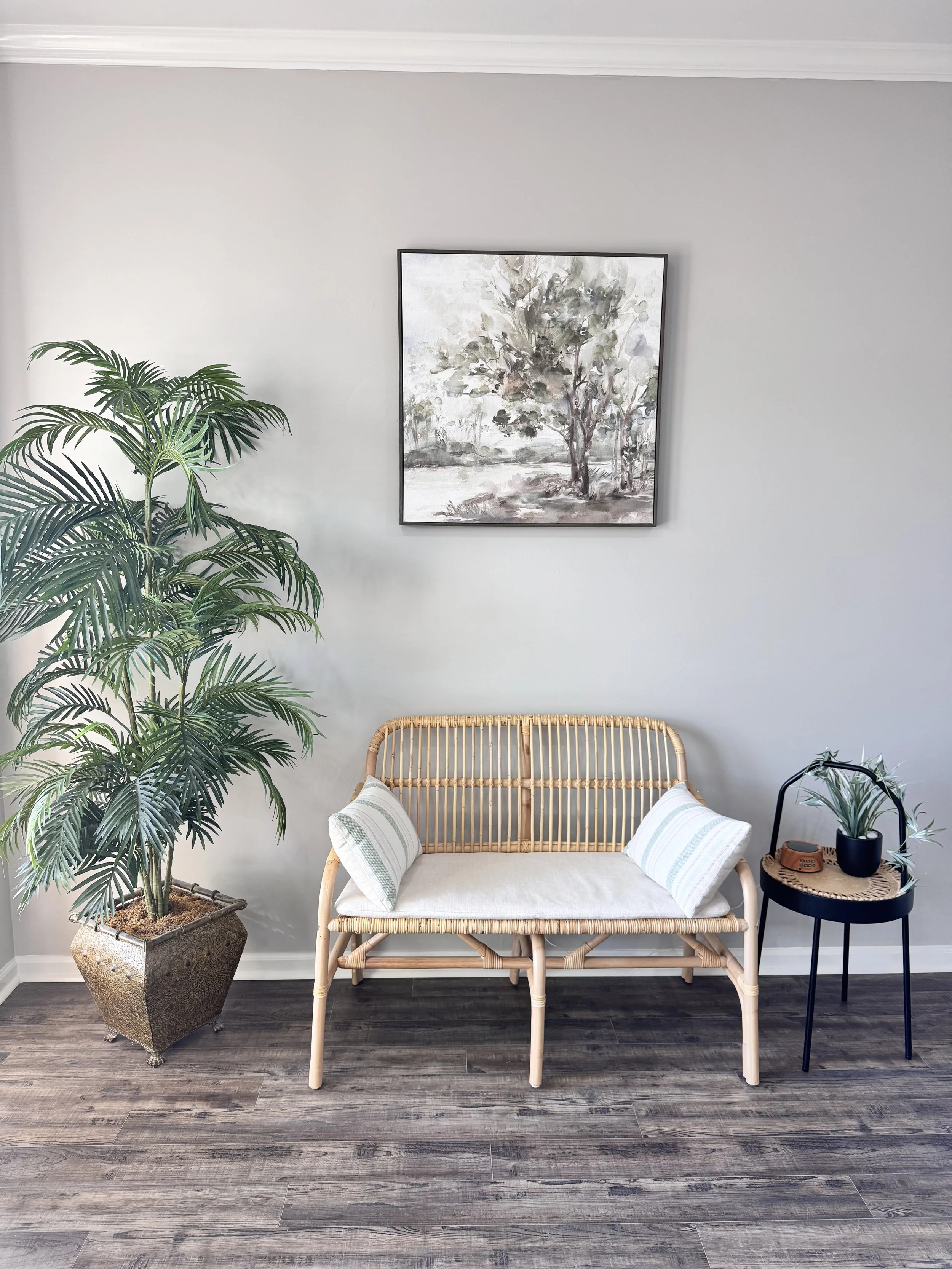 Minimalist living room interior with a bamboo bench, two cushions, a large potted plant, small round side table with a plant, and a landscape painting on a gray wall.