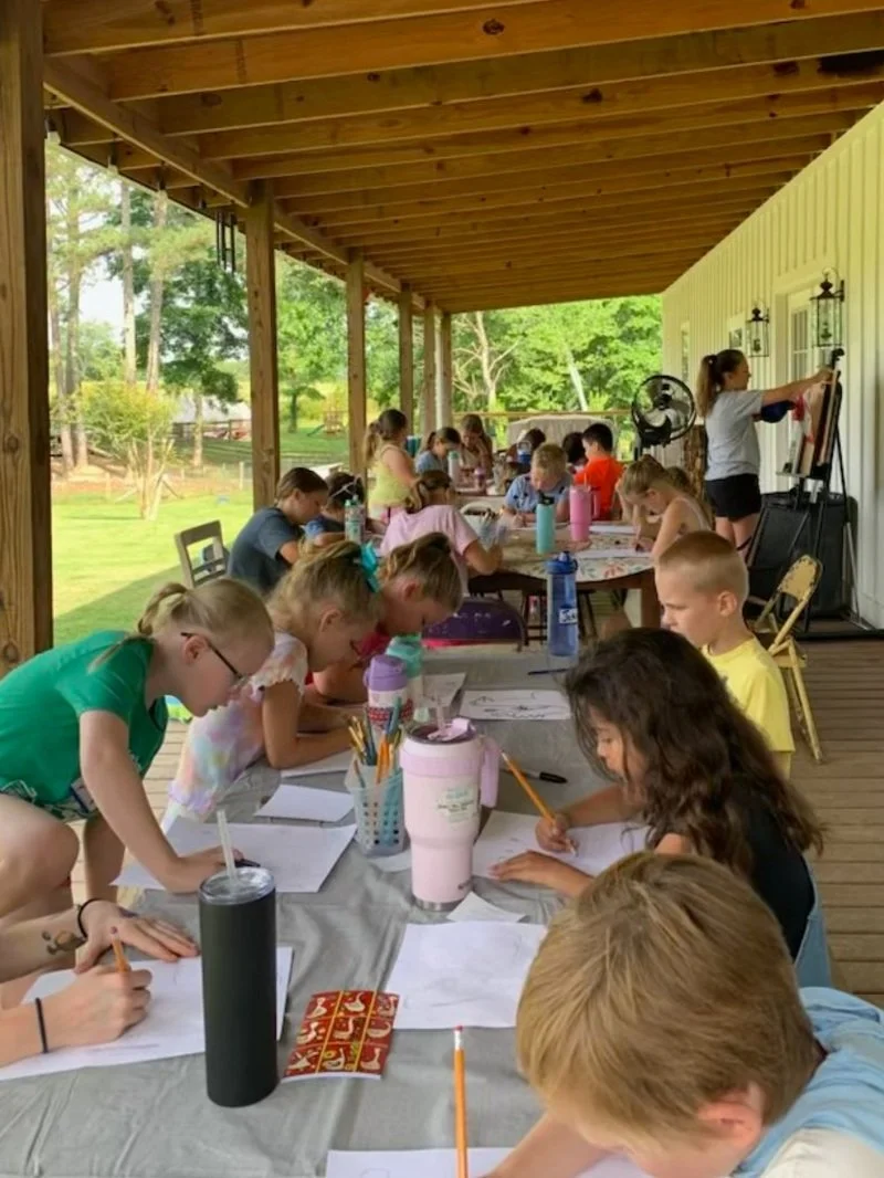 Children sitting at a long table drawing outdoors under a covered porch, with a woman instructing at a whiteboard.