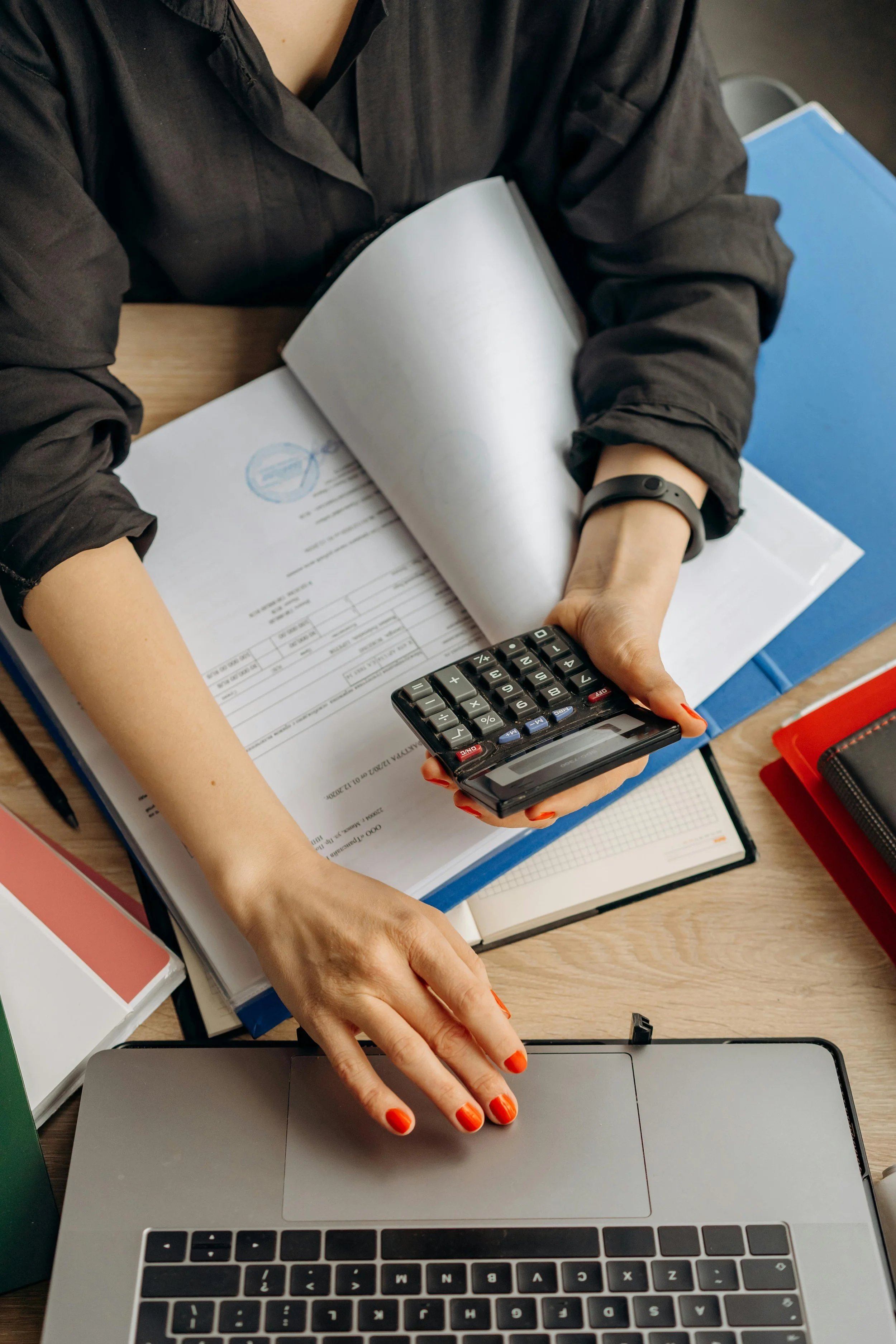 Top-down view of a person with red painted nails using a calculator, laptop, and paperwork on a desk.