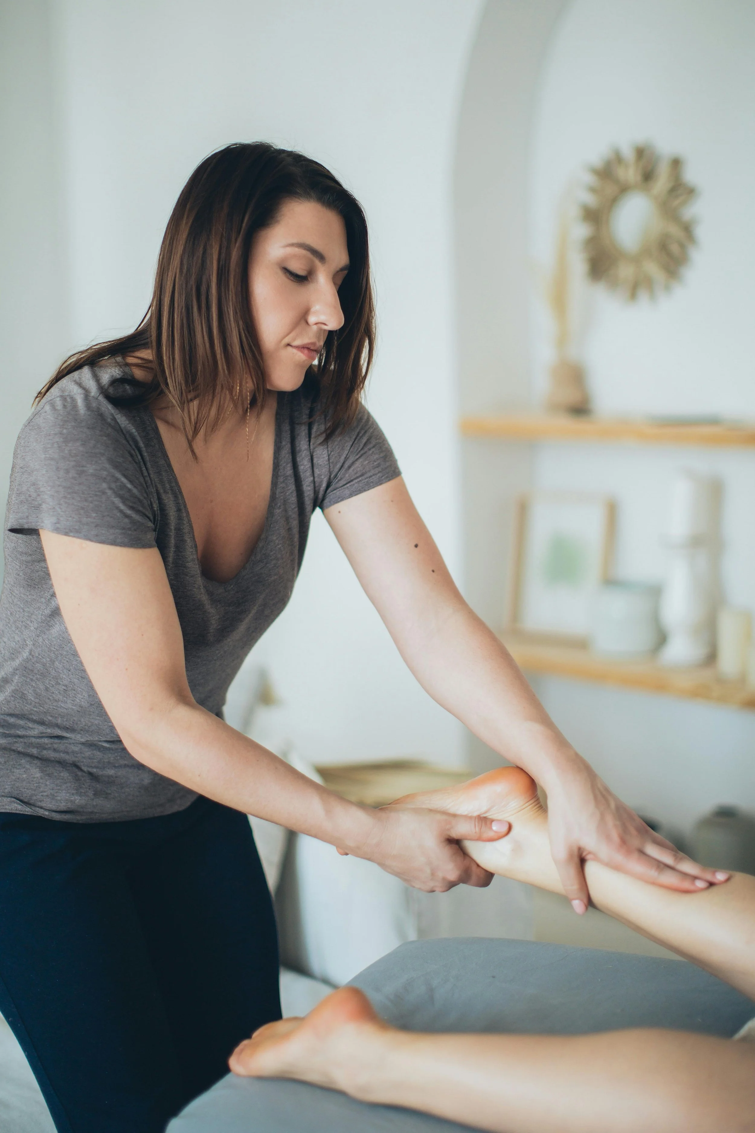 A woman in a gray shirt giving a foot massage to another person lying on a table in a spa or massage therapy setting.