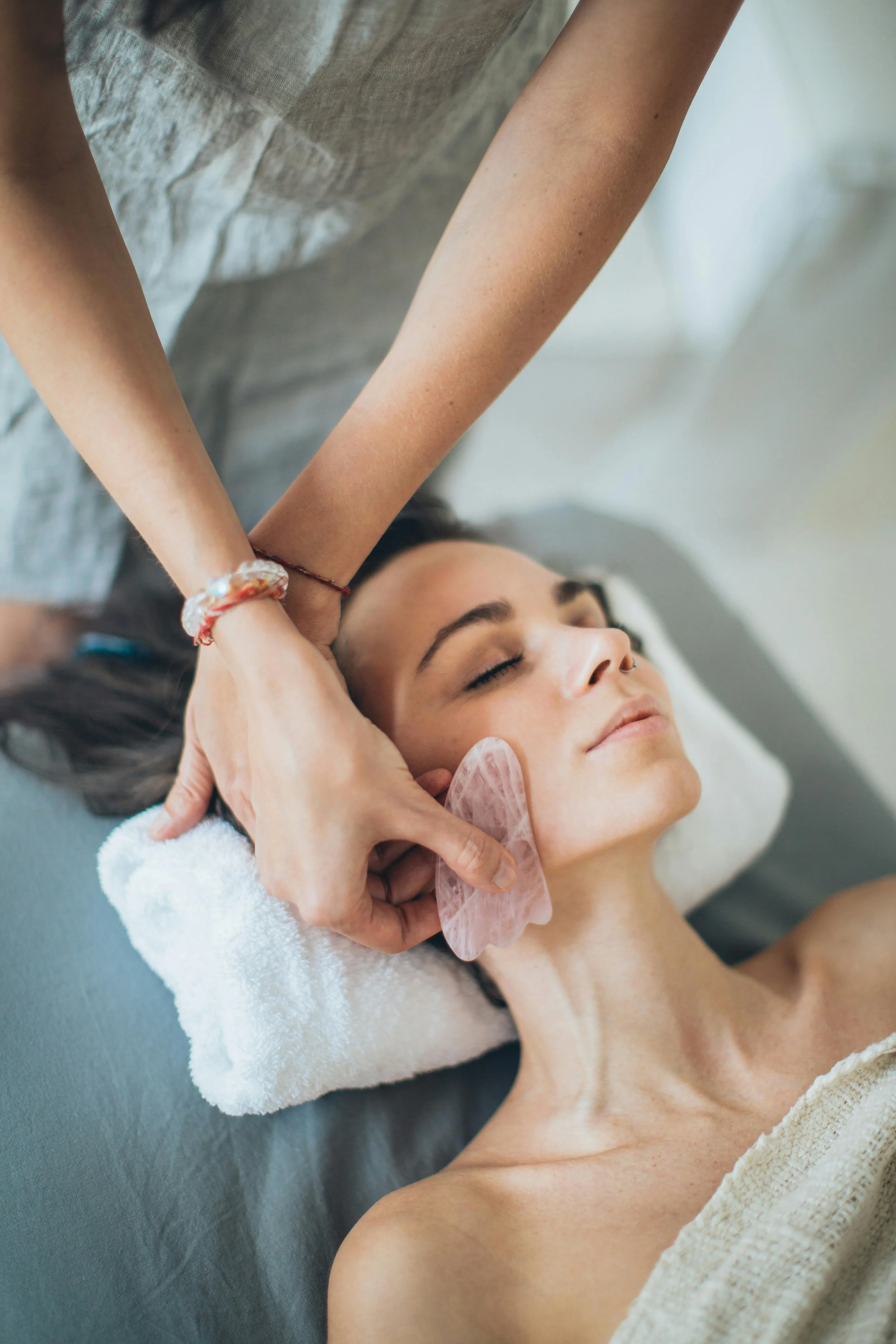 A woman receiving a facial treatment with a jade roller on her cheek, lying down with her eyes closed in a spa setting.