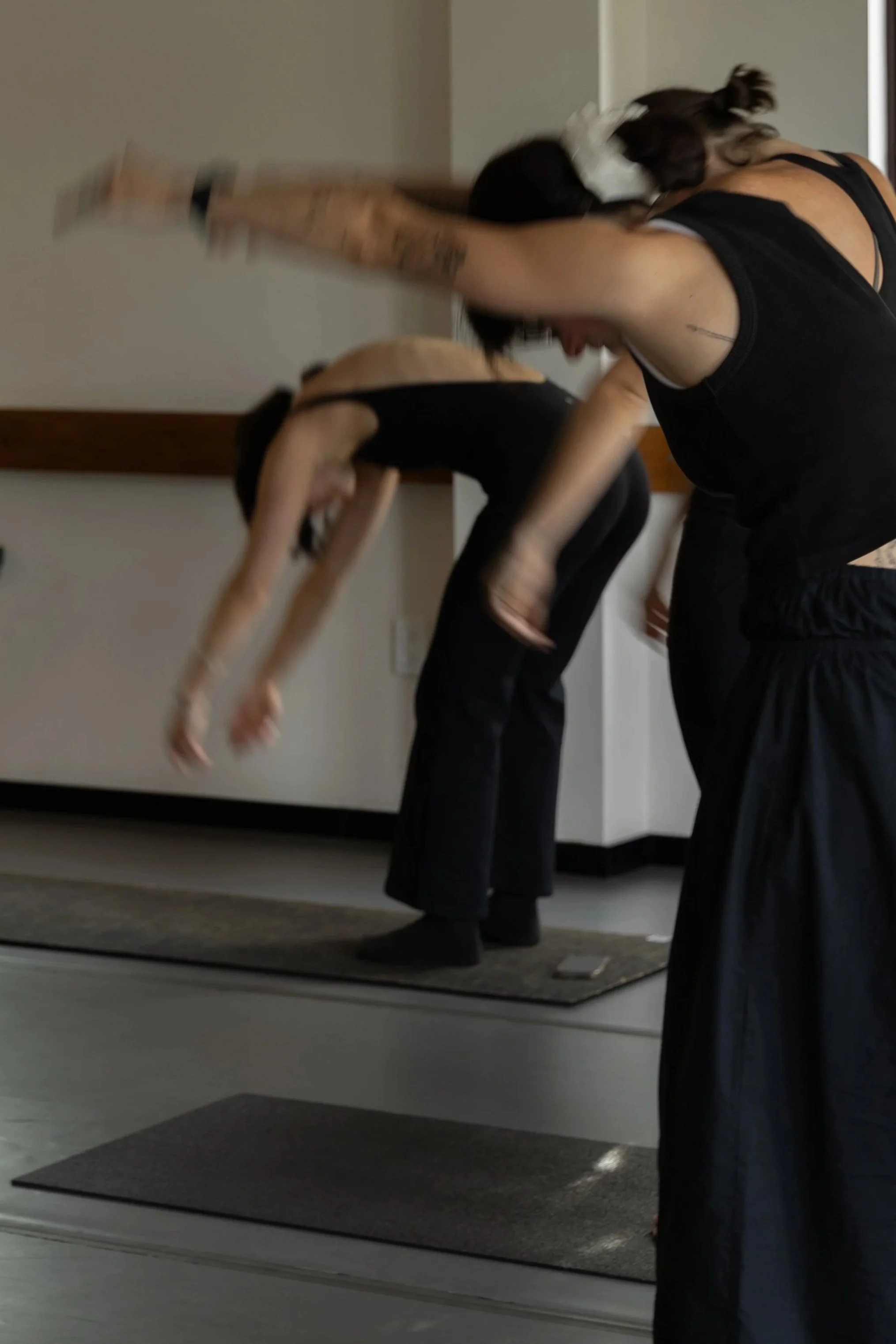 People in a pilates class lying on mats, raising their arms, led by an instructor in a room with a wooden floor and white walls.