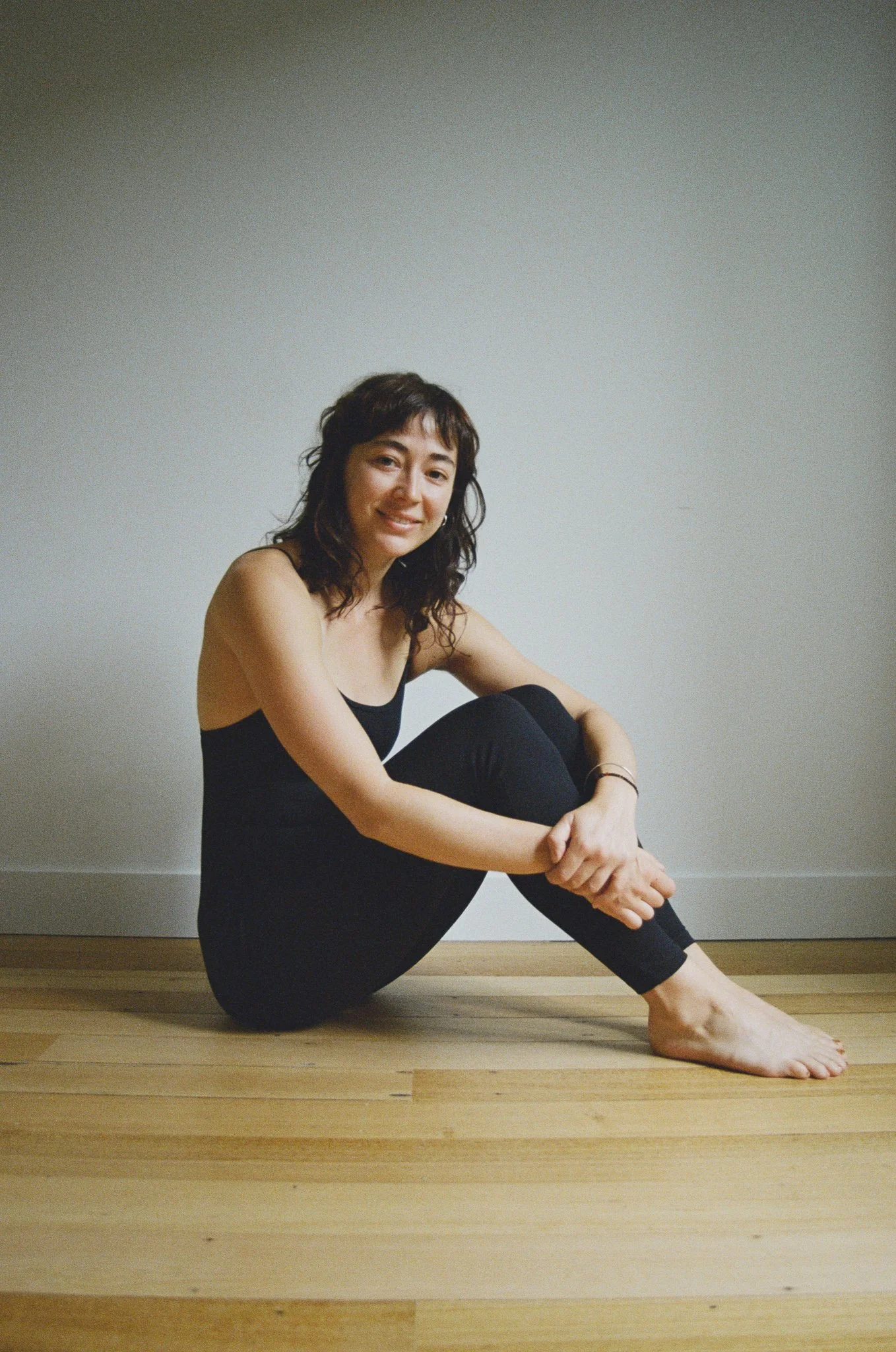Woman sitting on a wooden floor, wearing a black outfit, smiling, with her knees up, against a plain white wall.