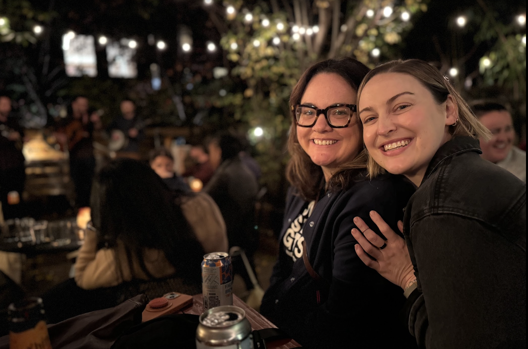 This is an image of Sam and Chelsey sitting at a picnic table in the backyard at Saturn Road - a cobble hill brooklyn coffee shop and wine bar. Sam wears black glases, a light jacket, and a t shirt. Chelsey wears a black denim jumpsuit