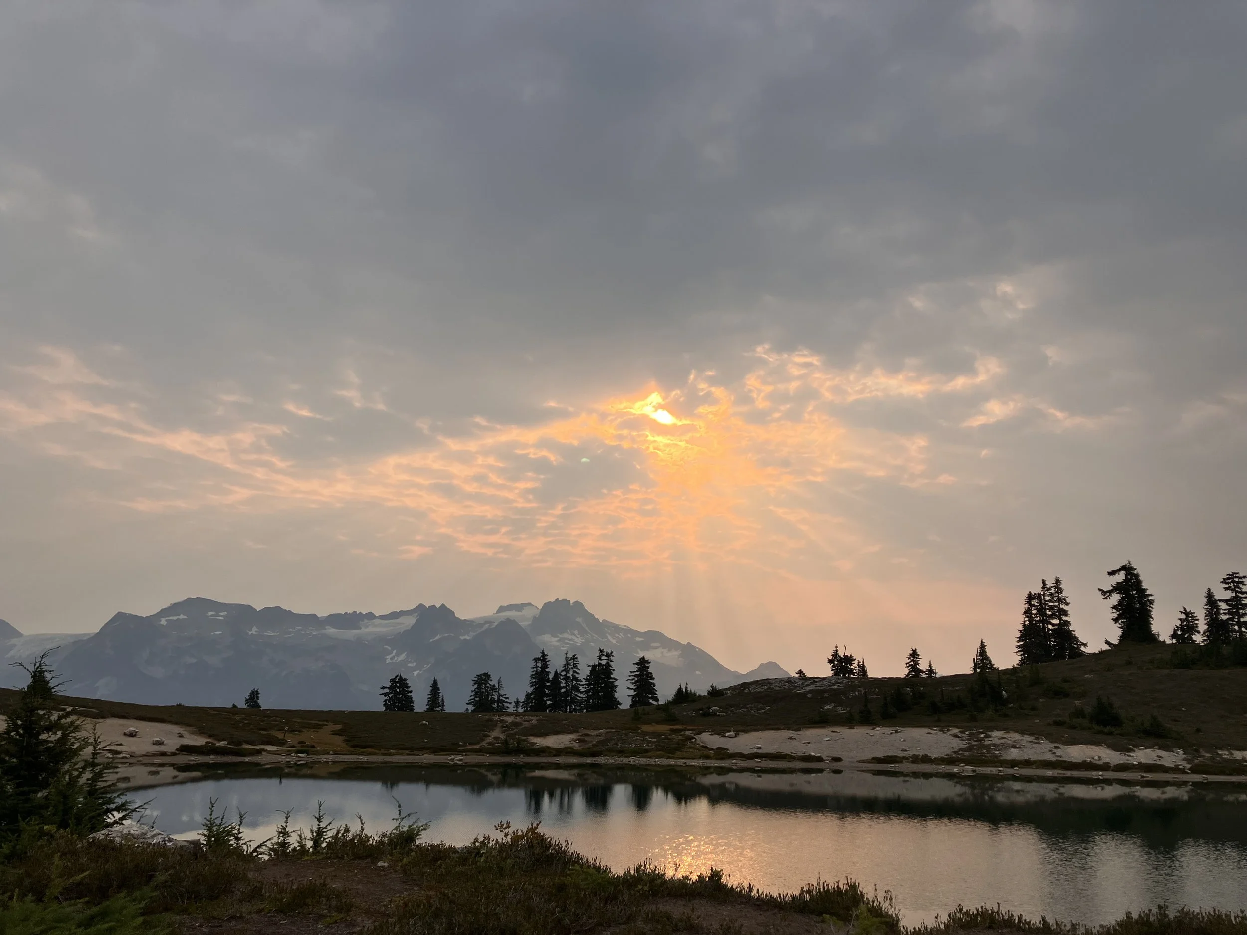 A serene landscape of a lake reflecting the sky and surrounded by pine trees, with mountains in the background and sun rays breaking through the clouds.