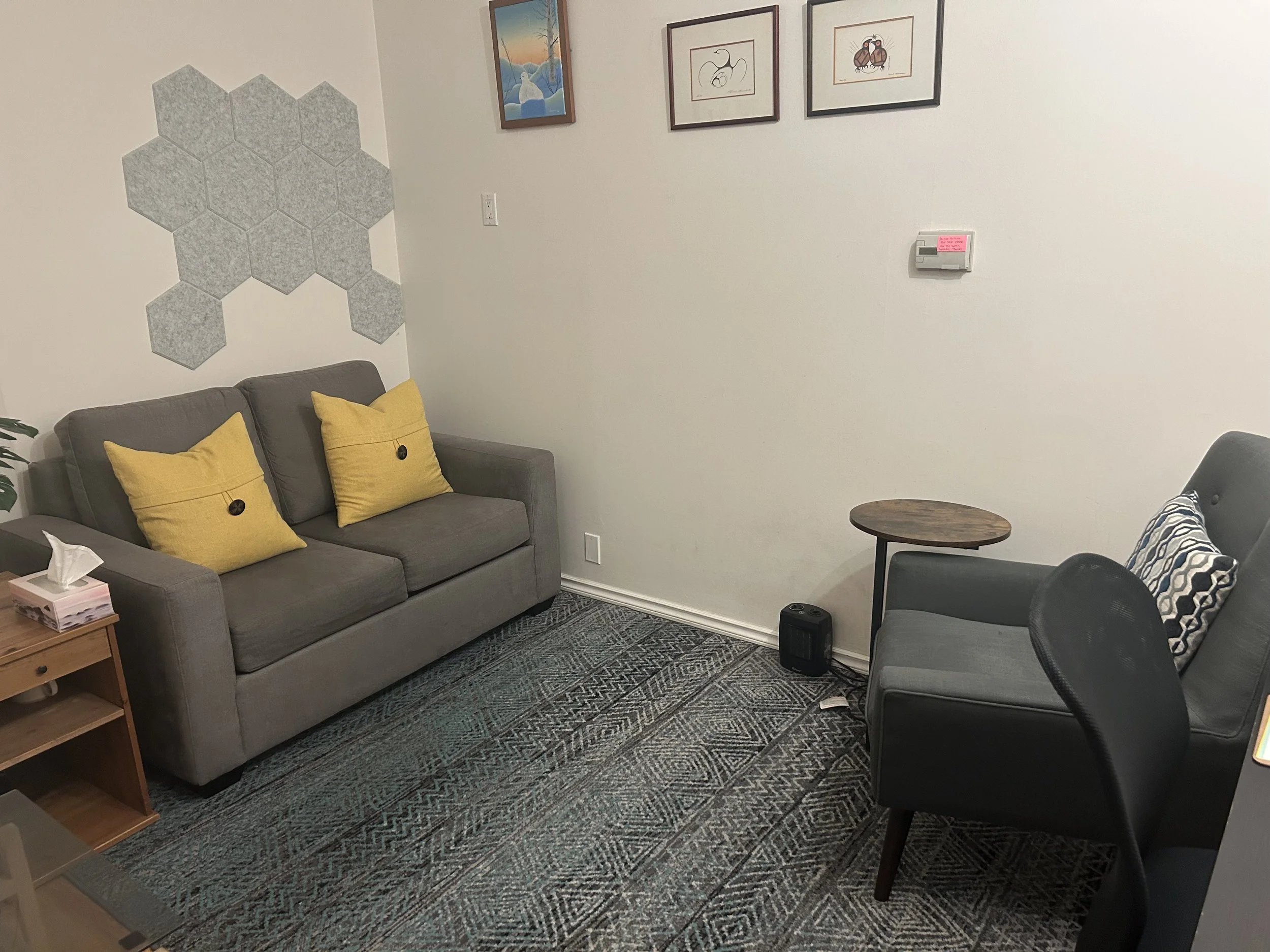 Small cozy office room with a gray sofa, two yellow pillows, a patterned armchair, a small round side table, and framed pictures on the wall.