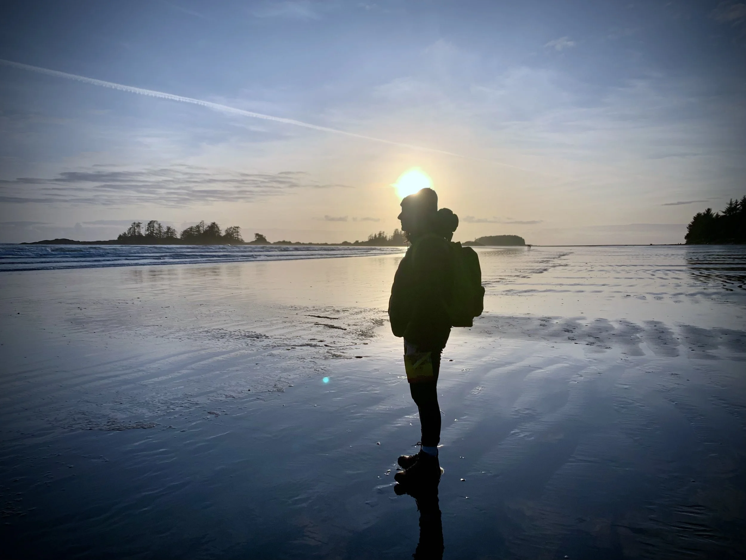 Silhouette of a person wearing a backpack standing on a beach at sunset, with the reflection on wet sand and distant trees on the horizon.