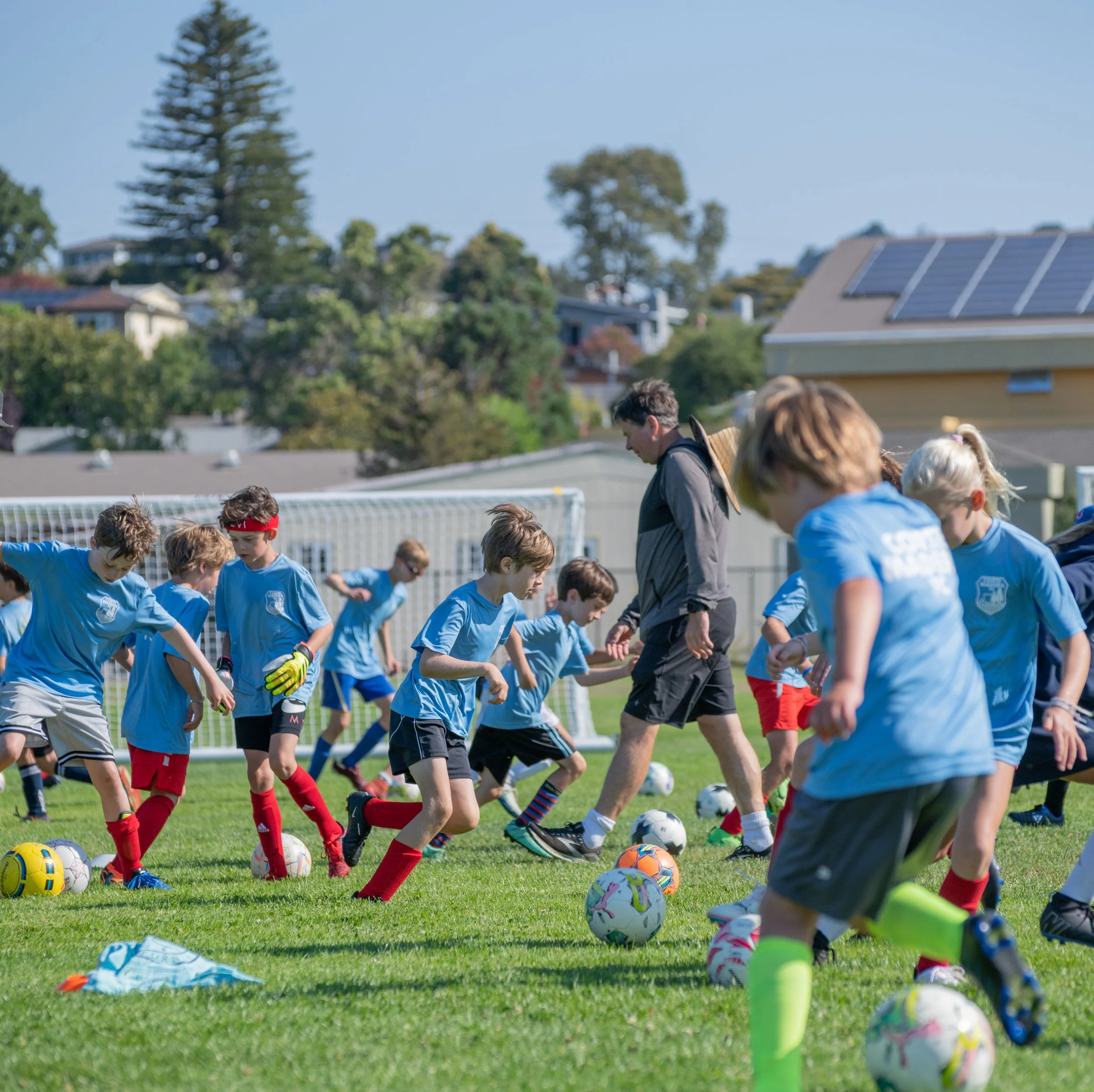 Children on a soccer field practicing with soccer balls, wearing blue jerseys, coached by an adult.