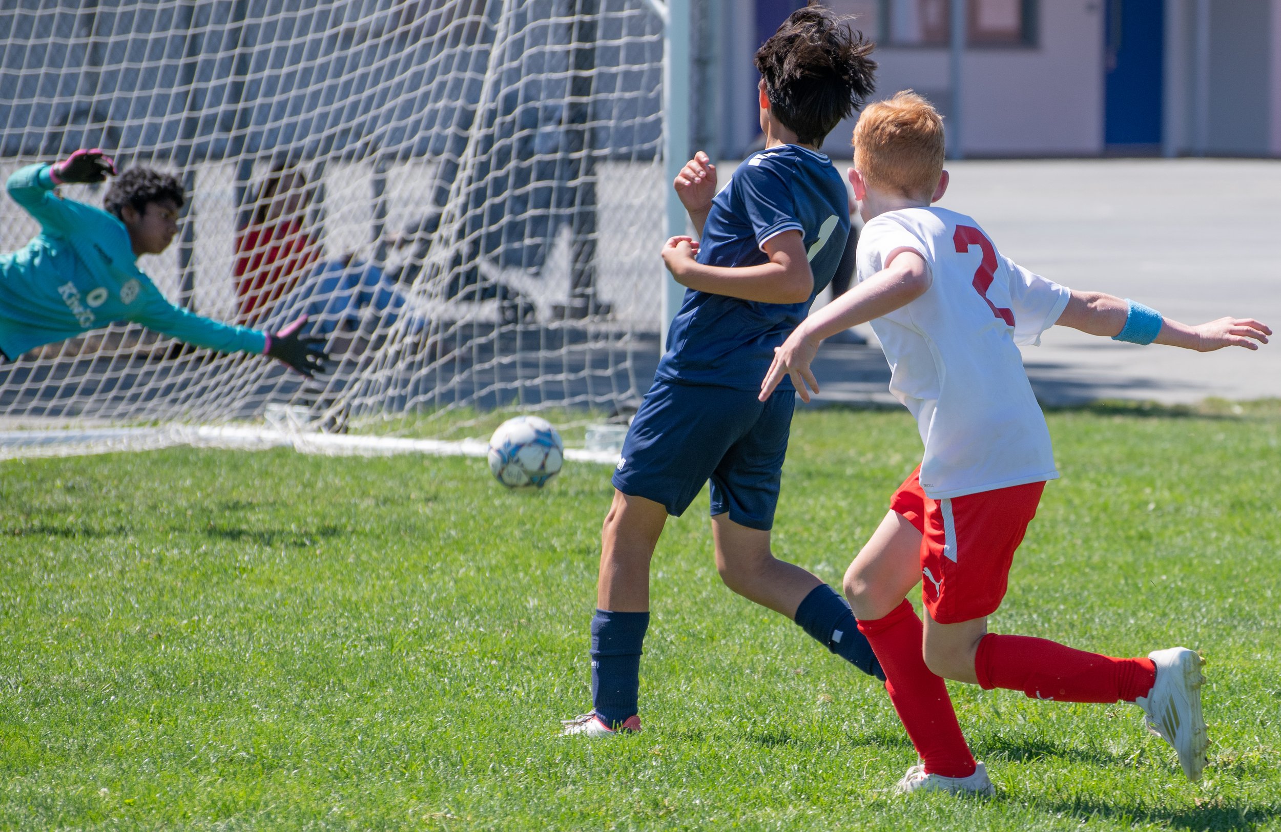 Two young soccer players, one in a blue uniform and one in a white and red uniform, are running towards the goal with a soccer ball, while a goalkeeper in a teal uniform is diving to save the shot on goal.