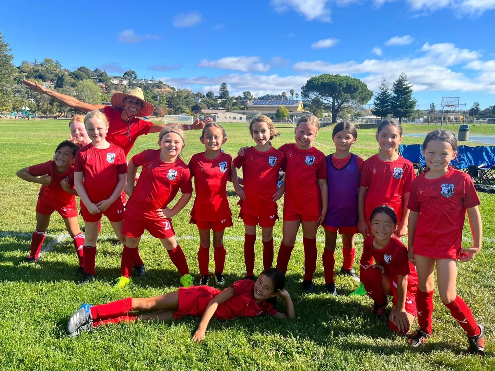 A group of young girls in red soccer uniforms and a coach in a sunhat posing on a soccer field with green grass, trees, and a blue sky with clouds in the background.