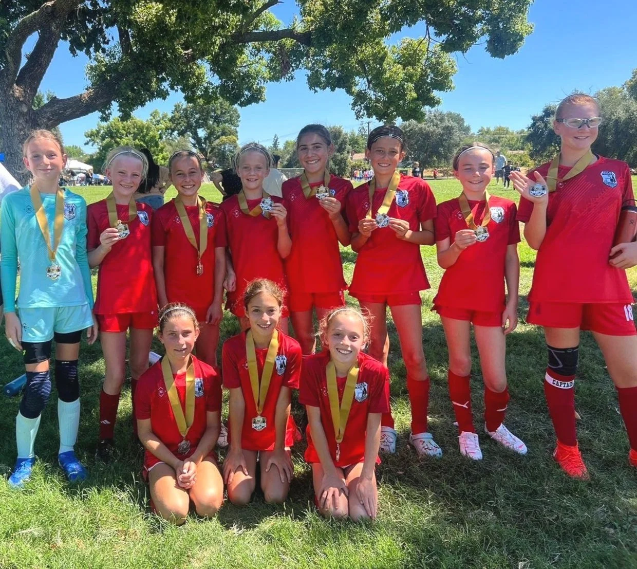 Group of young female soccer players in red uniforms holding medals, posing outdoors on a grassy field under a tree on a sunny day.