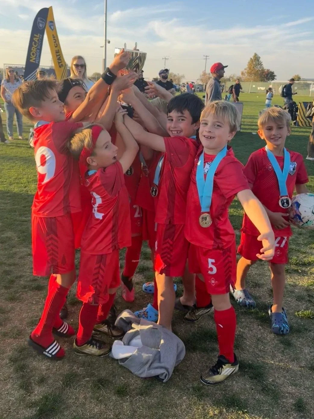 Children in red soccer uniforms celebrating with a trophy on a grassy field, wearing medals around their necks, with a background of people, flags, and a clear sky.